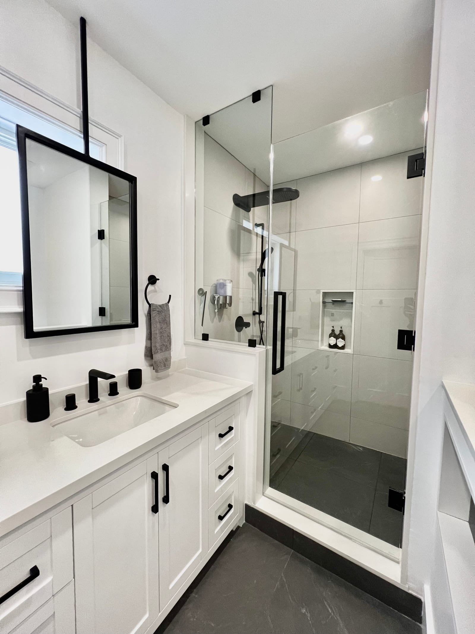 A white vanity with black fittings, a ceiling-hung black mirror and a glass shower with a niche