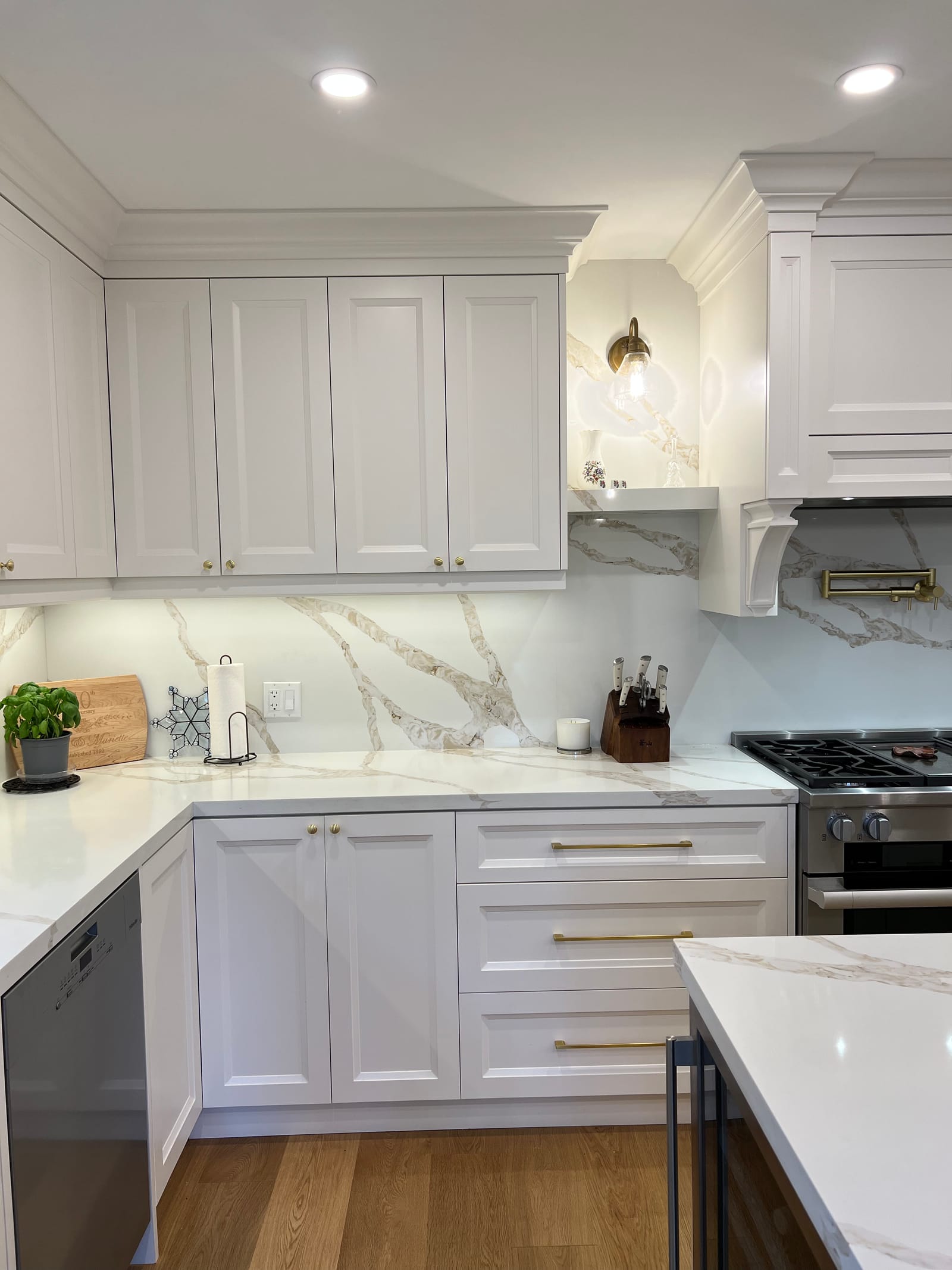 A white L-shaped run with a slab backsplash, a brass sconce over a floating shelf and a pot filler