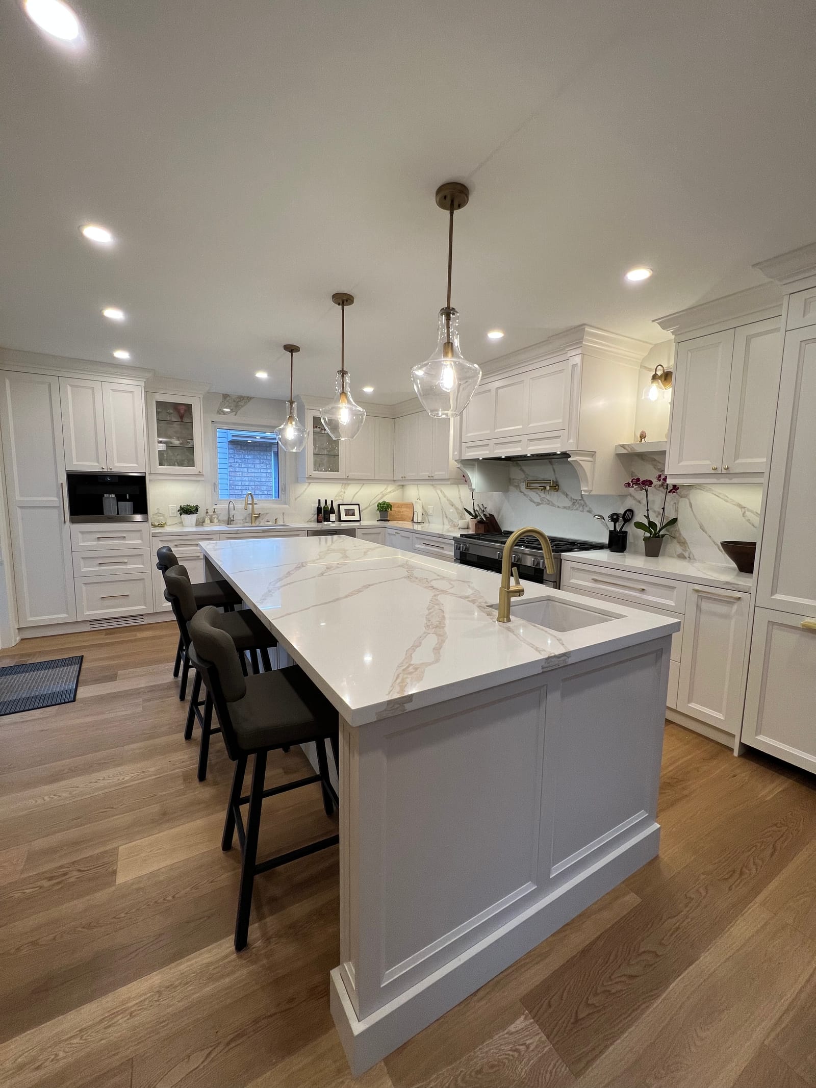 A white kitchen with a large island in gold-veined stone, three glass pendants and a range under a panelled hood
