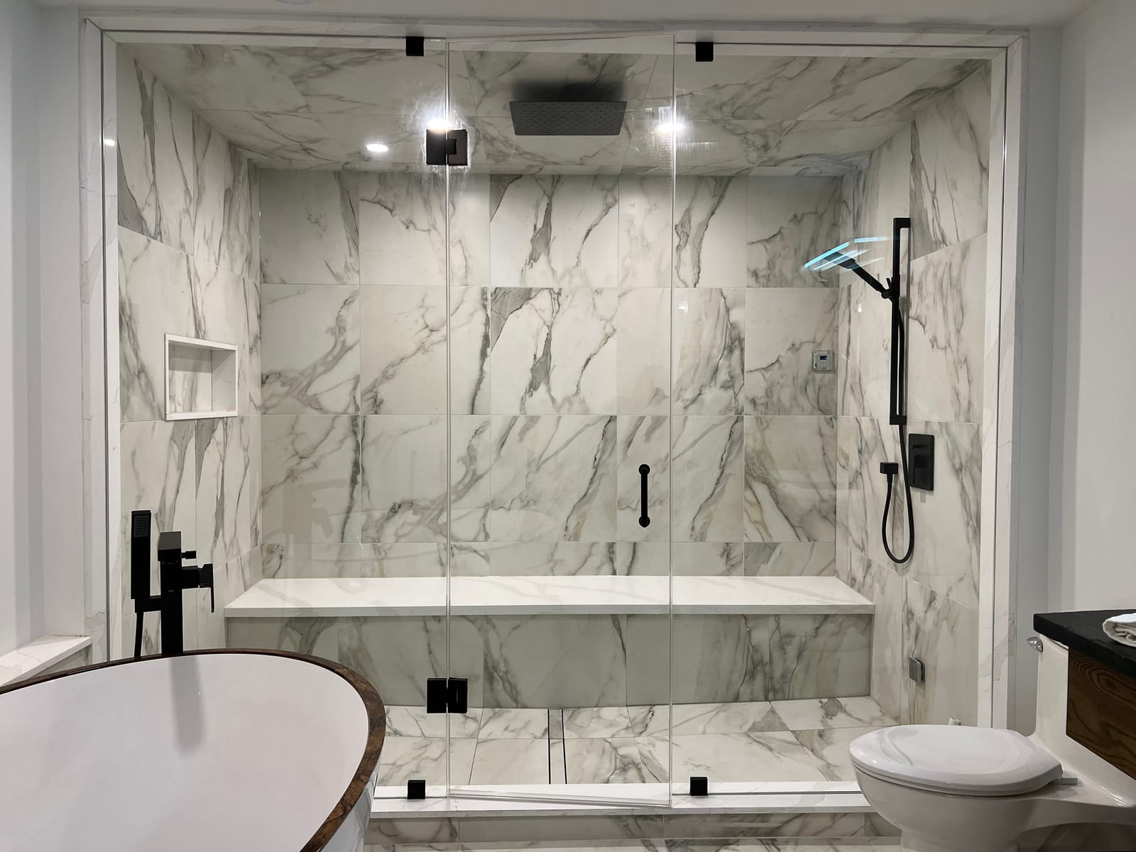 A large glass shower with a bench, clad in white marble-look slab with grey veining, beside a freestanding tub