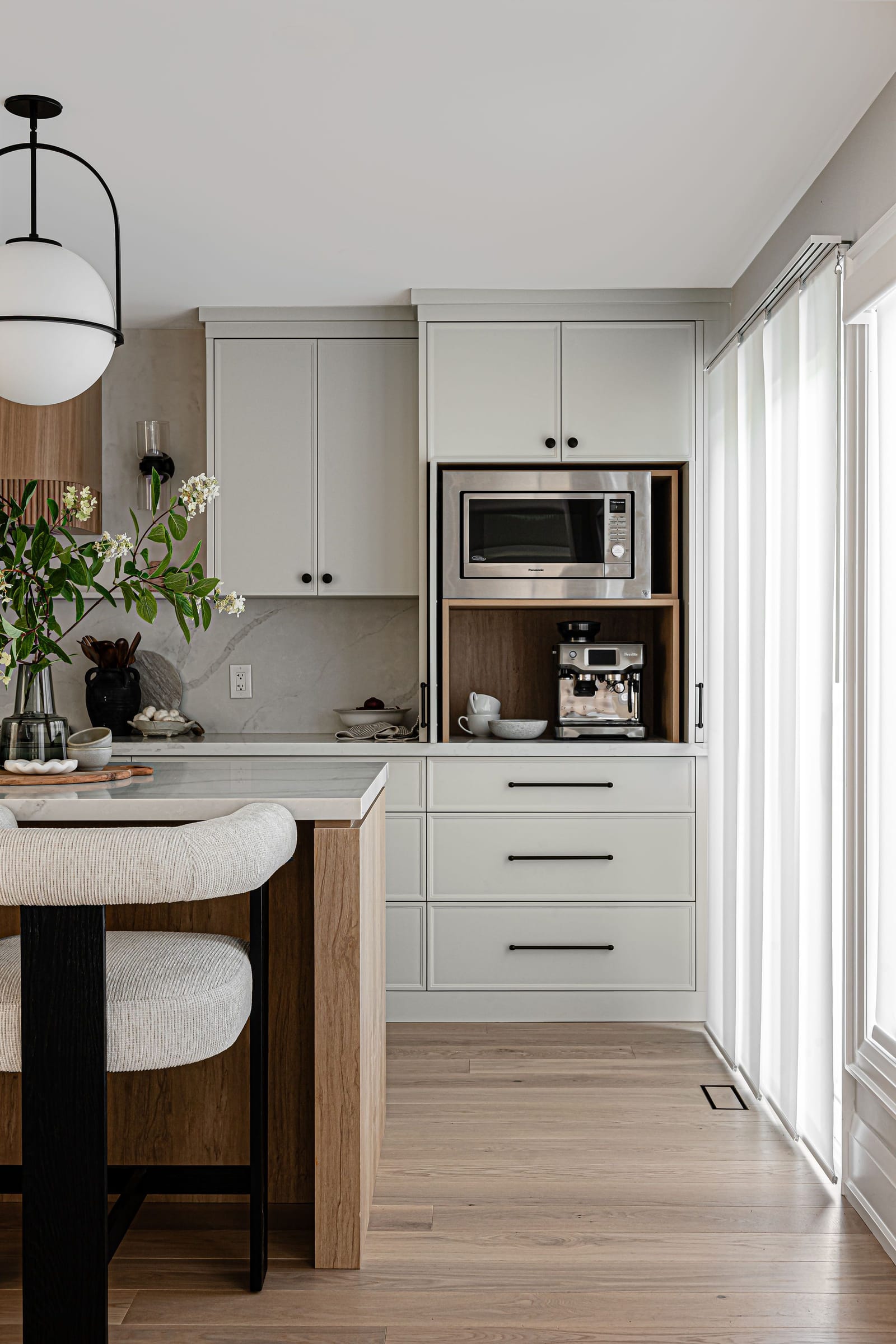 Tall white cabinets with an oak-lined niche holding a built-in microwave and an espresso machine, beside sheer-curtained glass doors