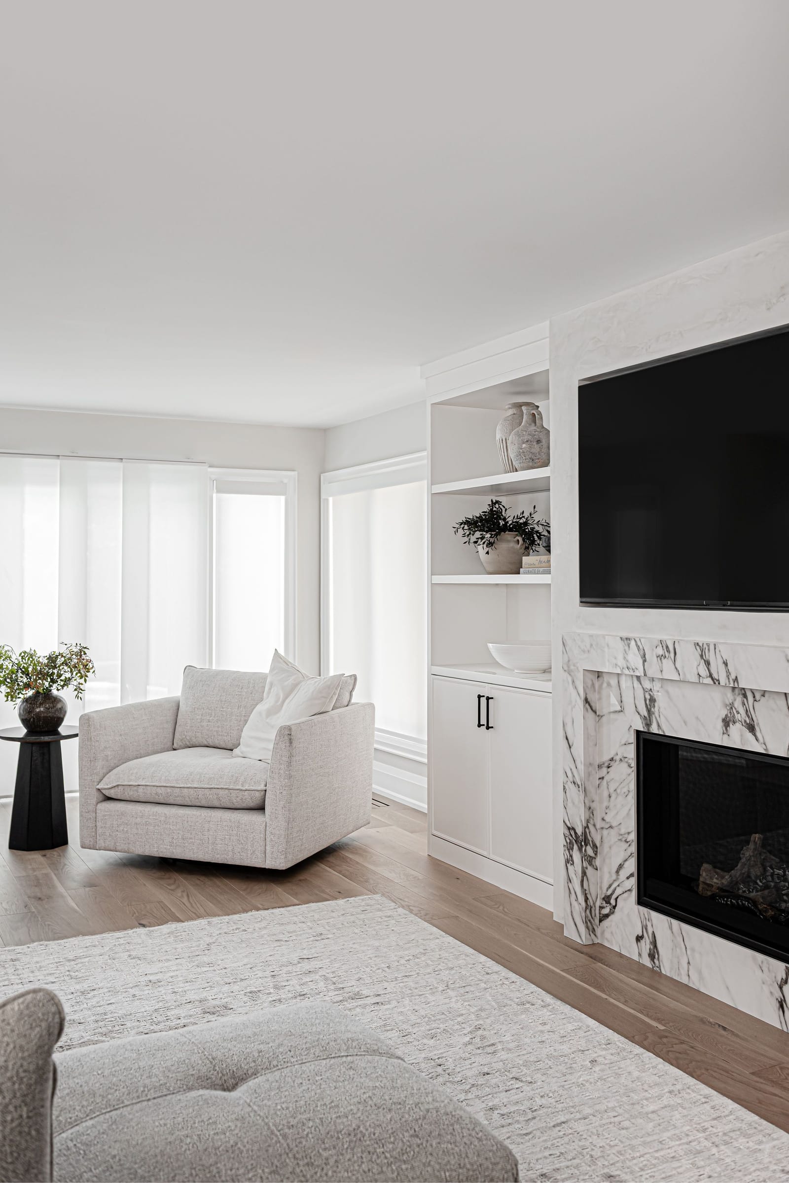 A gas fireplace faced in white stone with bold grey veining, with white built-in shelves and cabinets beside it and a swivel chair by the window