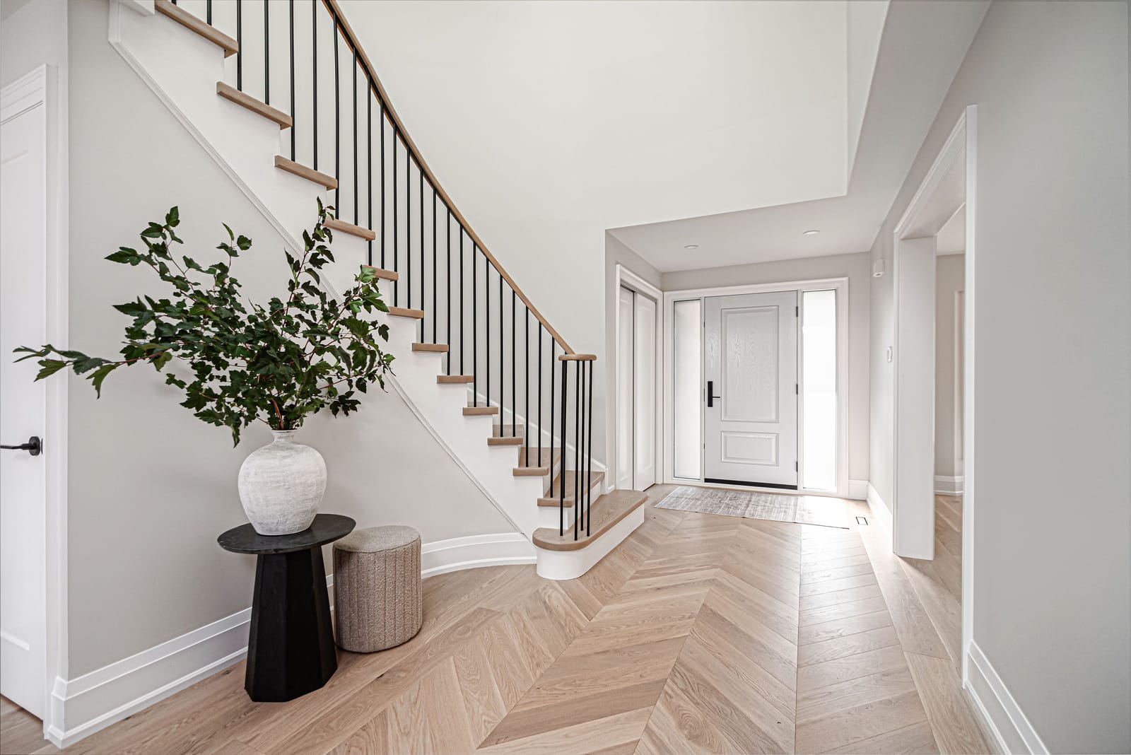 A foyer with a chevron white oak floor, a curved staircase with black spindles and the front door at the end of the hall