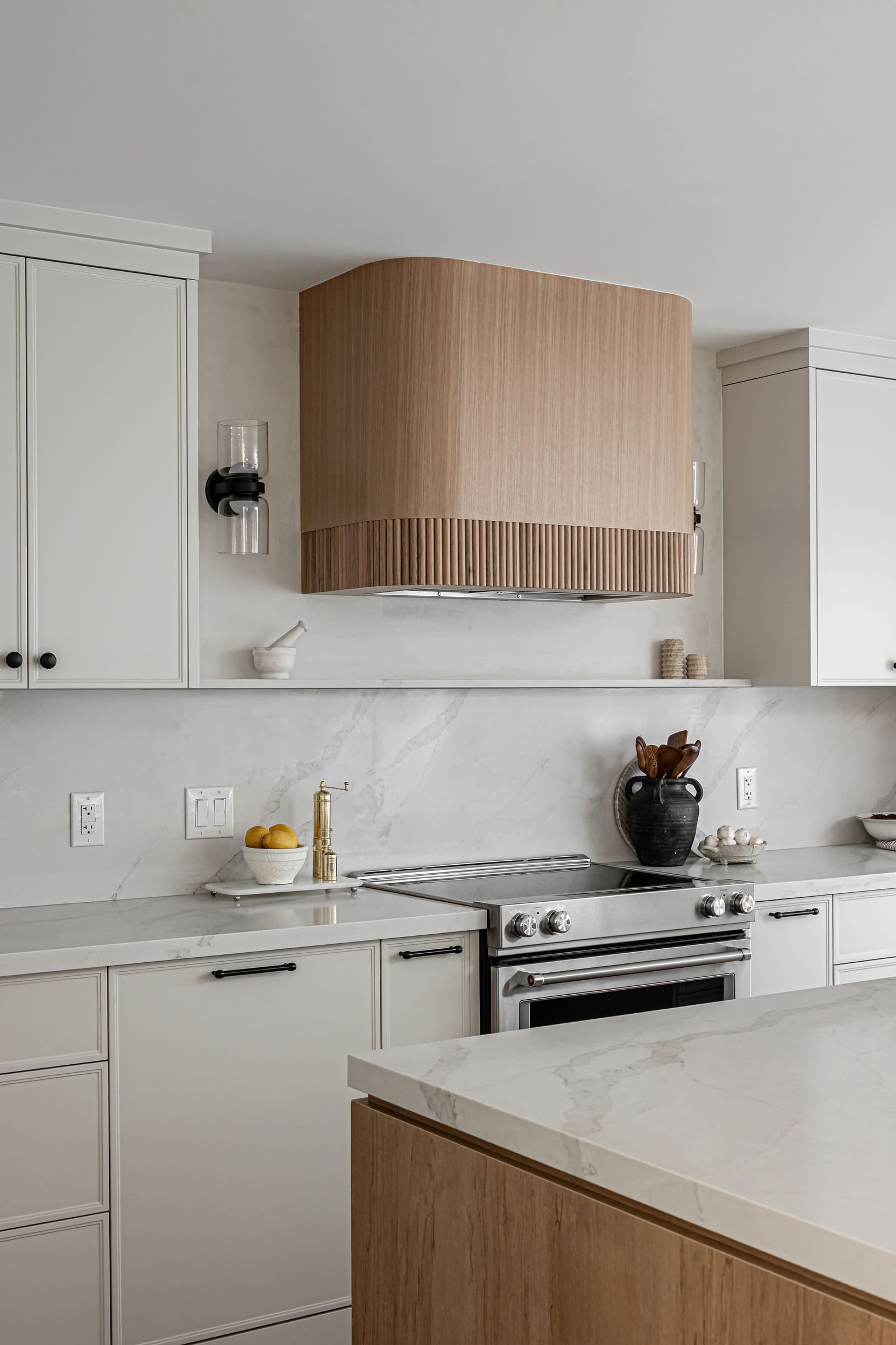 A curved range hood wrapped in fluted white oak above a stainless range, between white shaker cabinets and a white slab backsplash