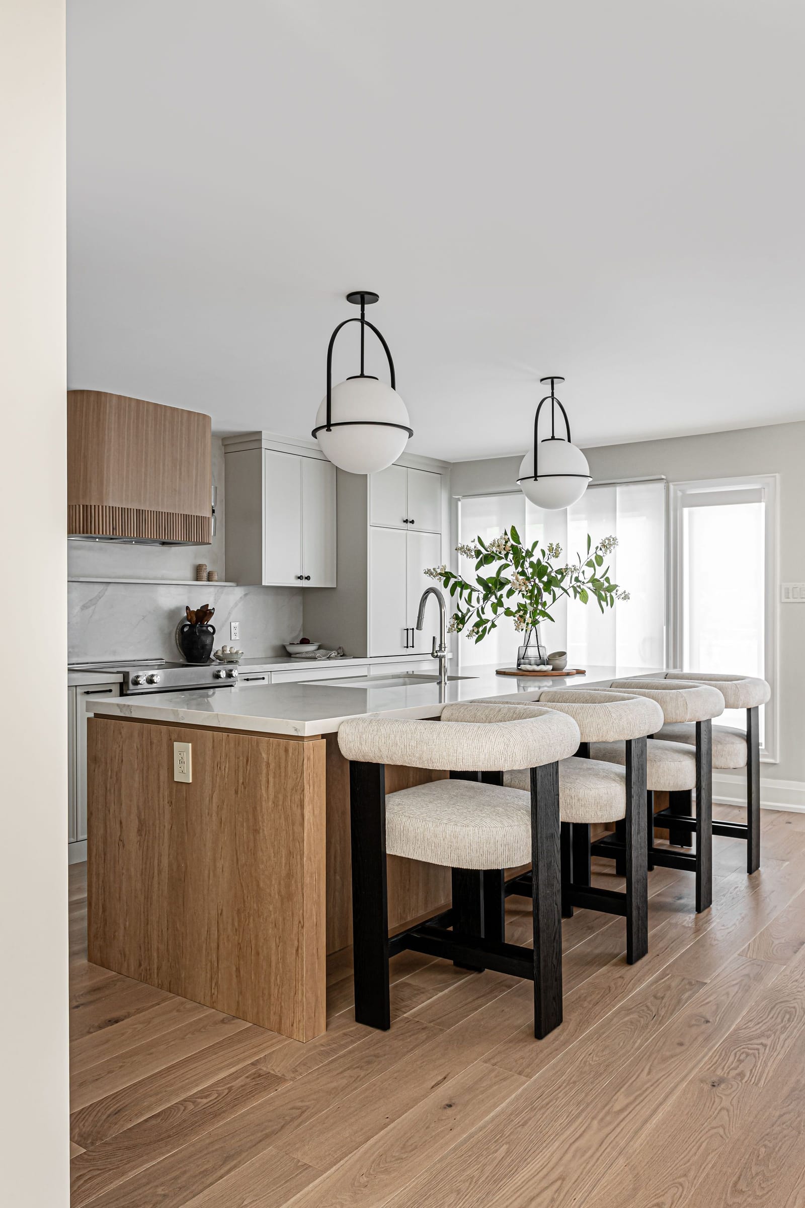 A white oak island with a white quartz top and four boucle stools with black legs, under two globe pendants