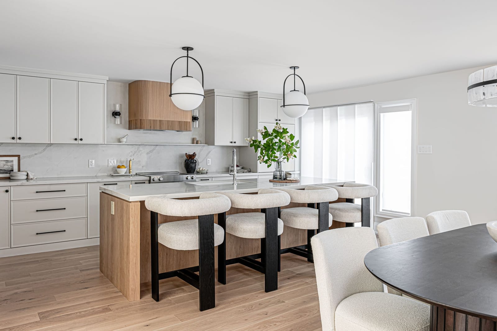 An open kitchen and dining area: a white oak island with four boucle stools, white cabinets, a fluted oak hood and an oval dining table