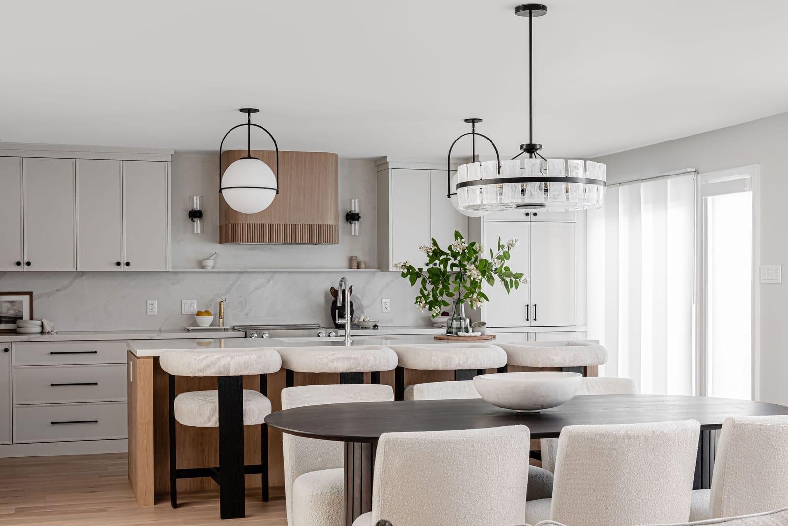 The kitchen wall of white cabinets with a fluted oak hood and slab backsplash, seen past an oval dining table and a crystal chandelier