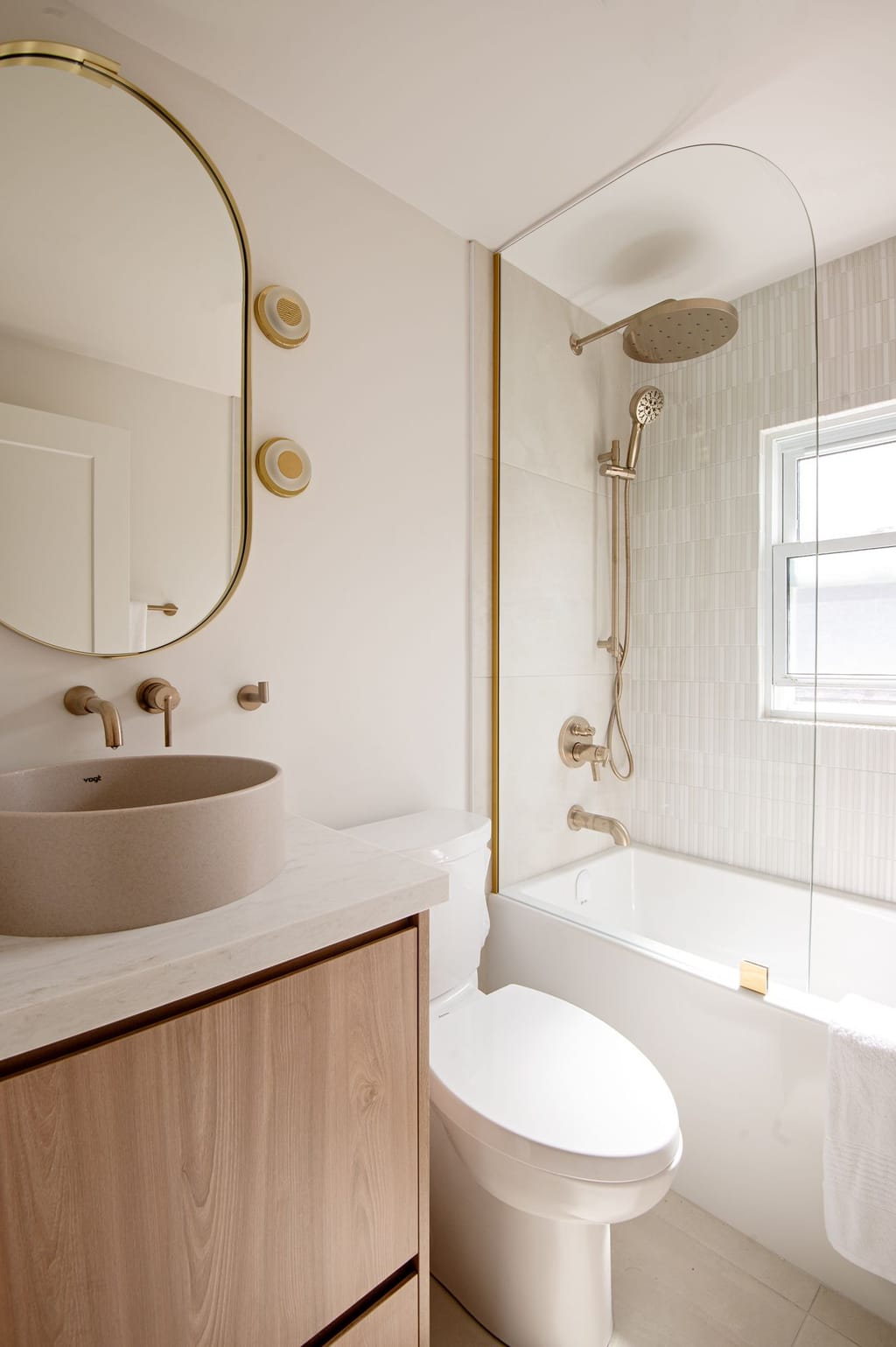 A bathroom with a stone vessel sink on an oak vanity, an oval brass mirror and a tub with a curved glass screen