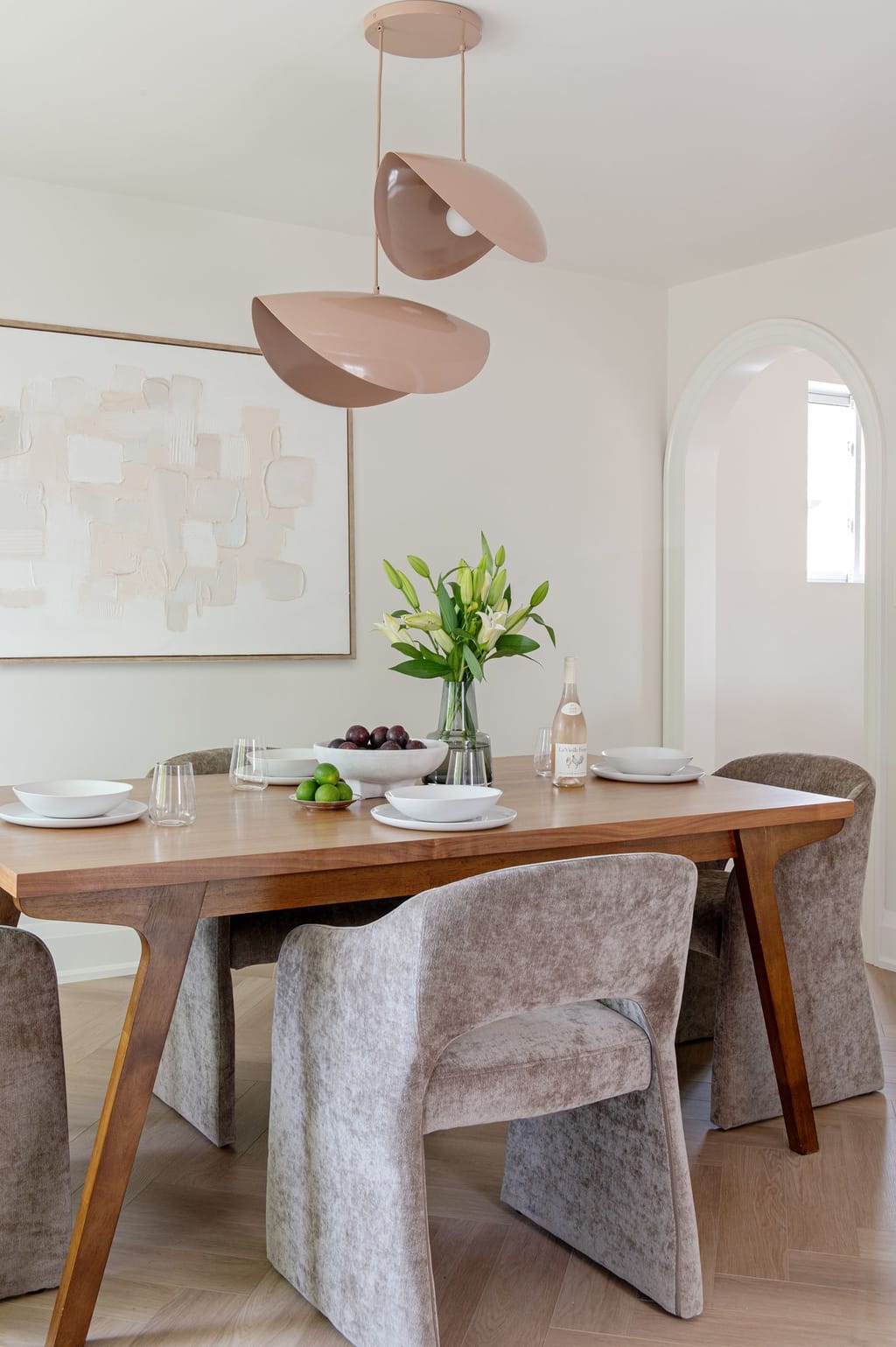 A walnut dining table with taupe velvet chairs under blush shell pendants, with an arched doorway behind