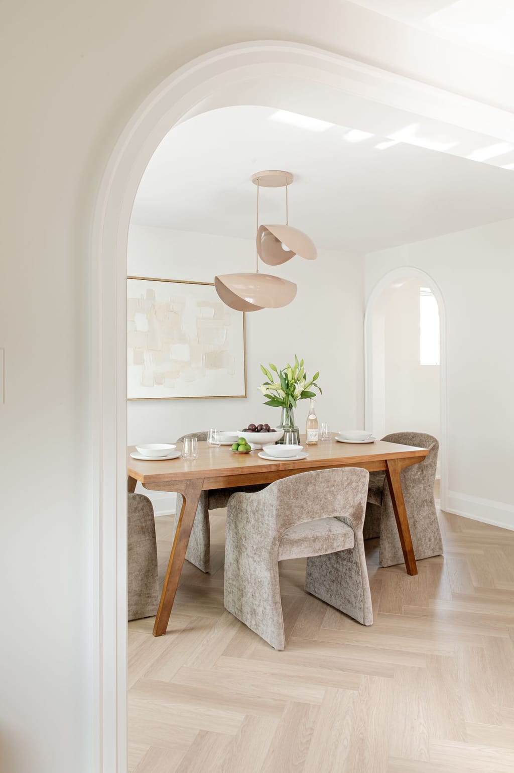 A wide arched opening into a dining room with a walnut table, velvet chairs and a pair of blush shell pendants