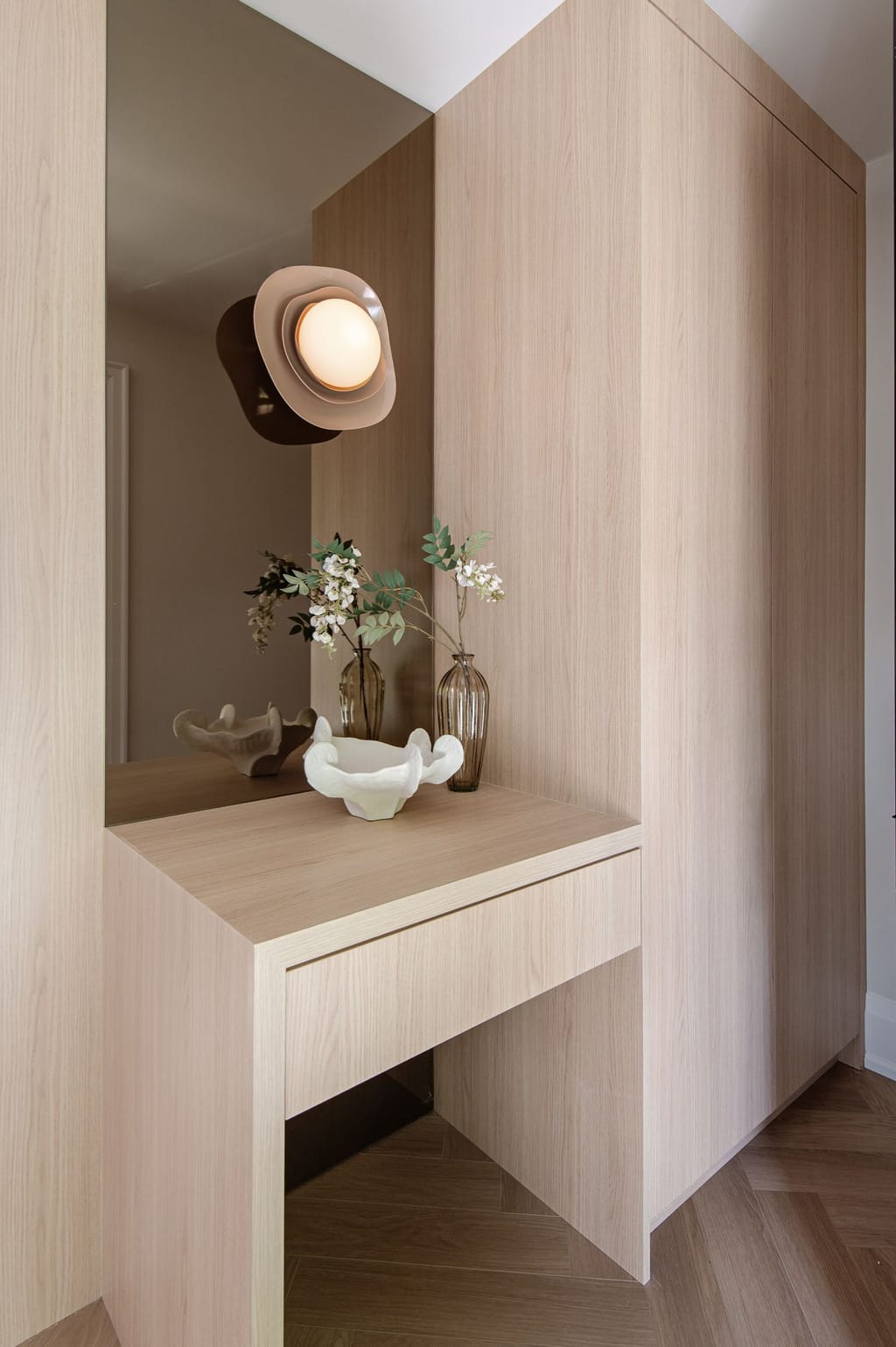 A pale oak dressing table built into a wall of oak wardrobes, with a mirror and a sculptural wall light