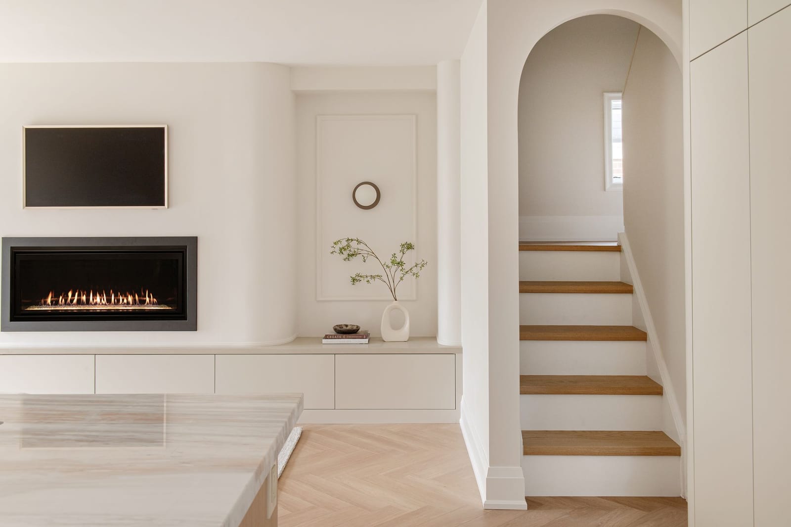 A linear fireplace in a curved plaster wall over a long low cabinet, beside an arched opening to an oak stair