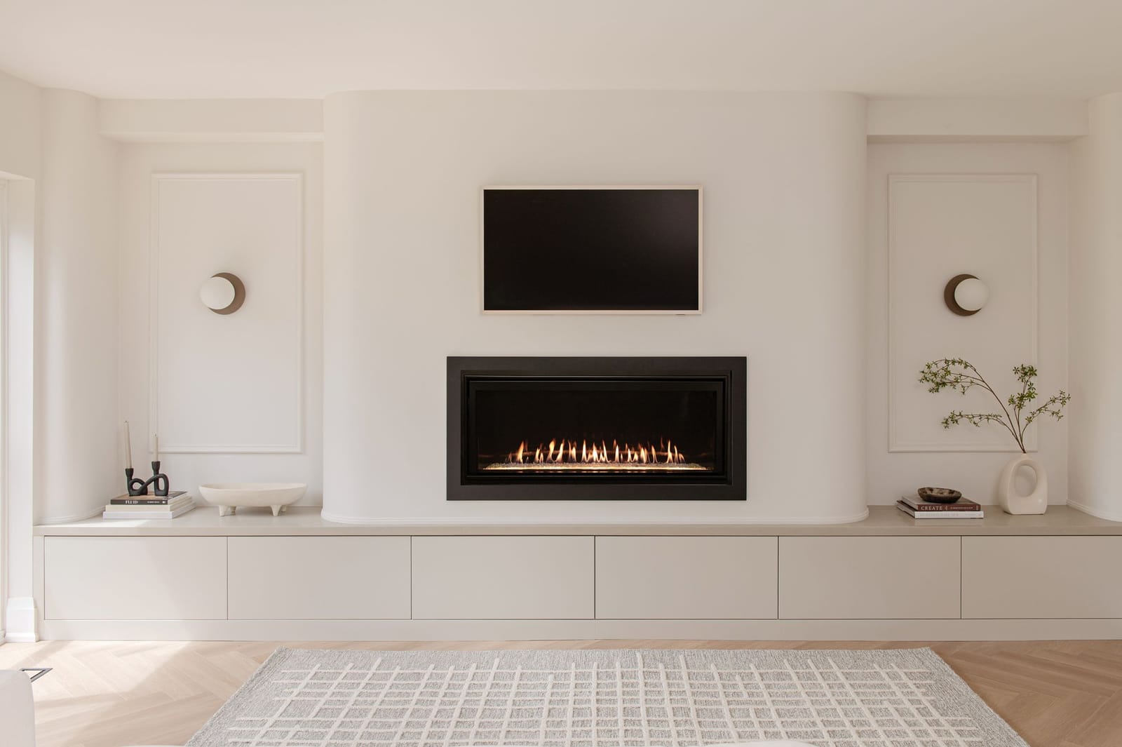 A linear fireplace and a TV set in a curved plaster wall, over a long low cream cabinet, with a panel and a round sconce on each side