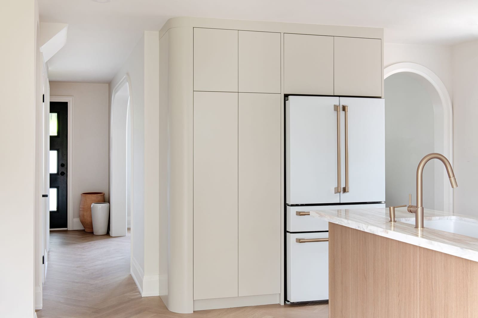 A rounded cream cabinet wall around a white fridge with bronze handles, beside an arched doorway
