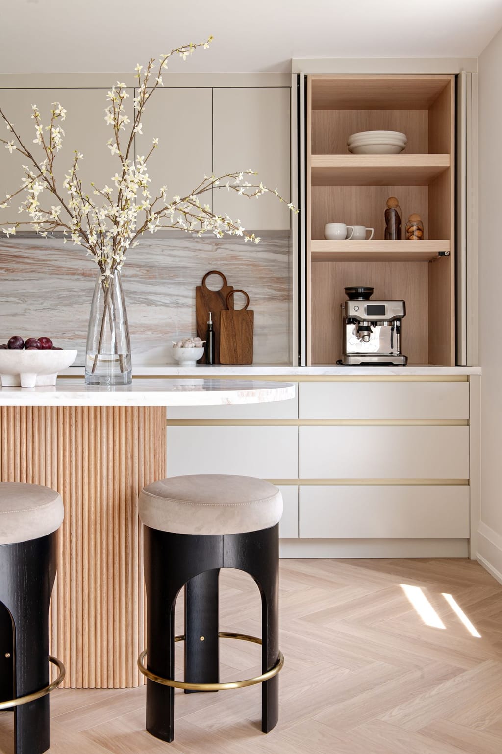 The rounded end of a fluted oak island with black stools, in front of an oak-lined coffee cabinet with pocket doors