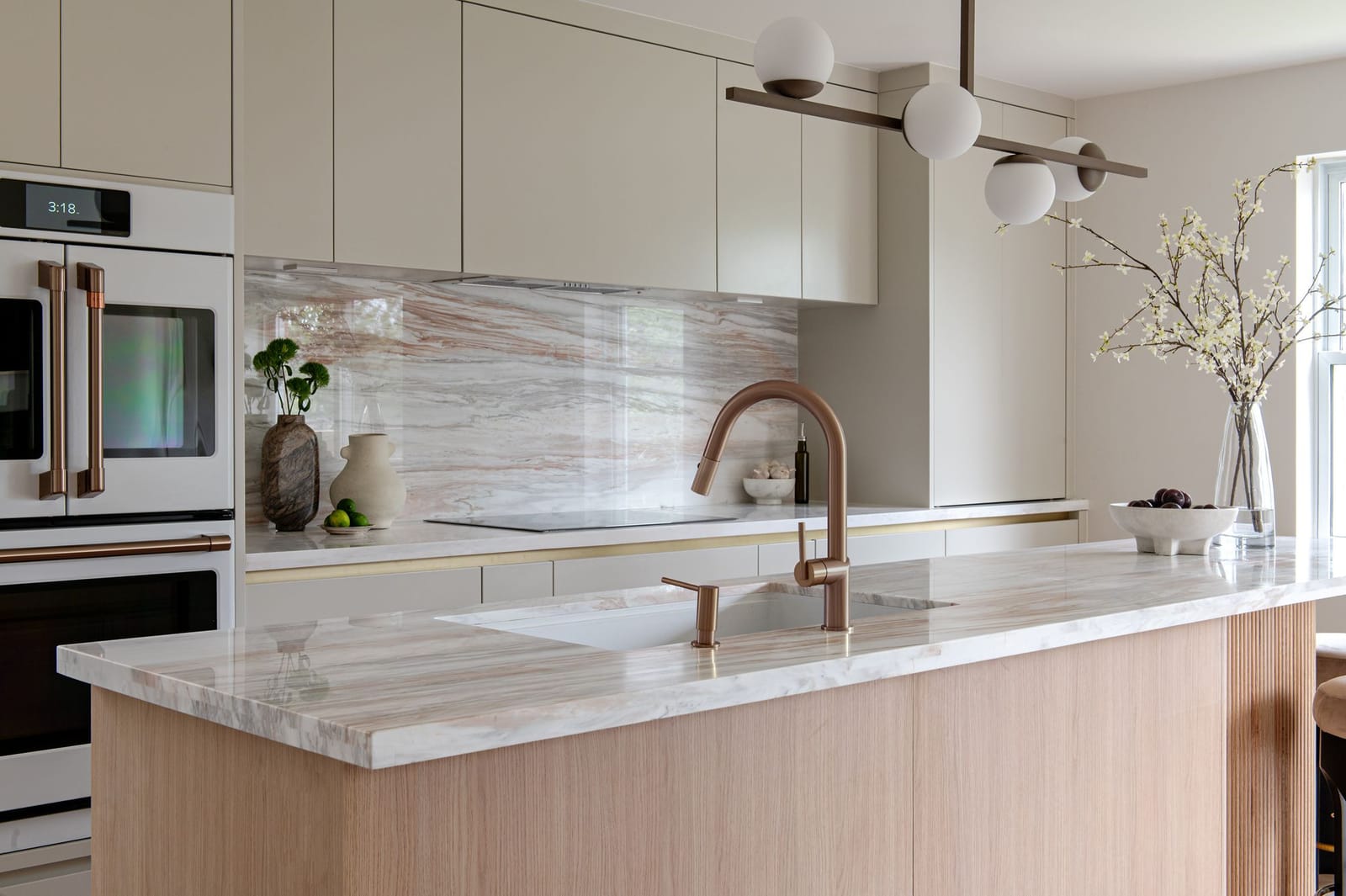 A pink-veined stone island top with a copper faucet, in front of white wall ovens and cream slab cabinets
