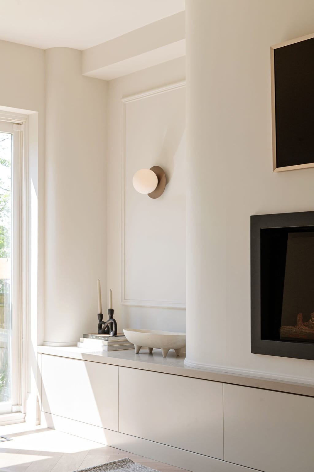 A sunlit living room corner with a long low cream cabinet, a picture-frame panel wall, a round sconce and a fireplace