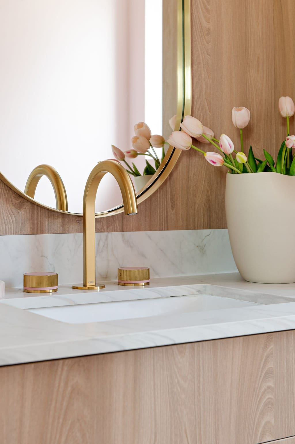 A brushed gold faucet on a marble counter under an oval brass mirror, with pink tulips against oak panelling