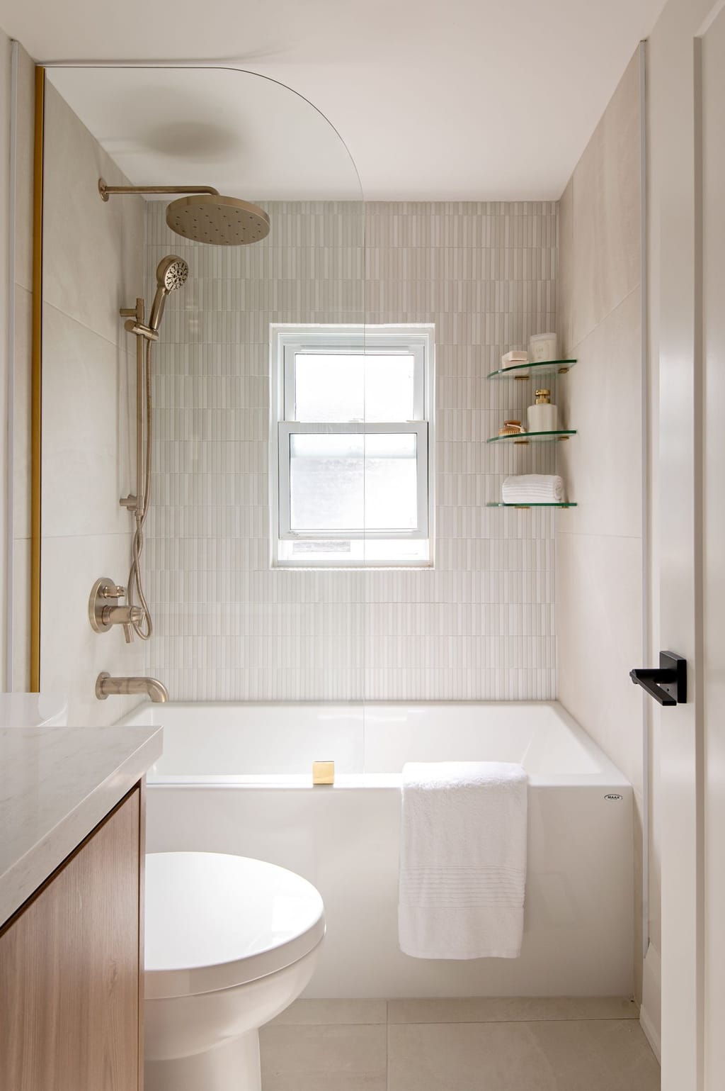 A tub alcove in thin white vertical tile with a window, glass corner shelves and brushed bronze fittings