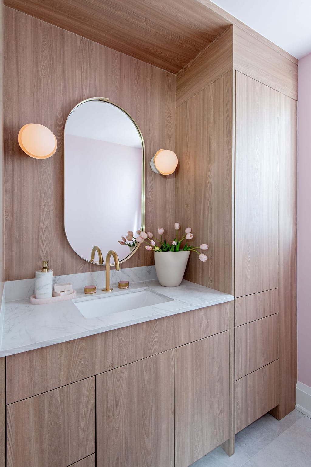A wood-panelled vanity wall that wraps onto the ceiling, with an oval brass mirror, two globe sconces, a white veined counter and a tall cabinet, against pink walls