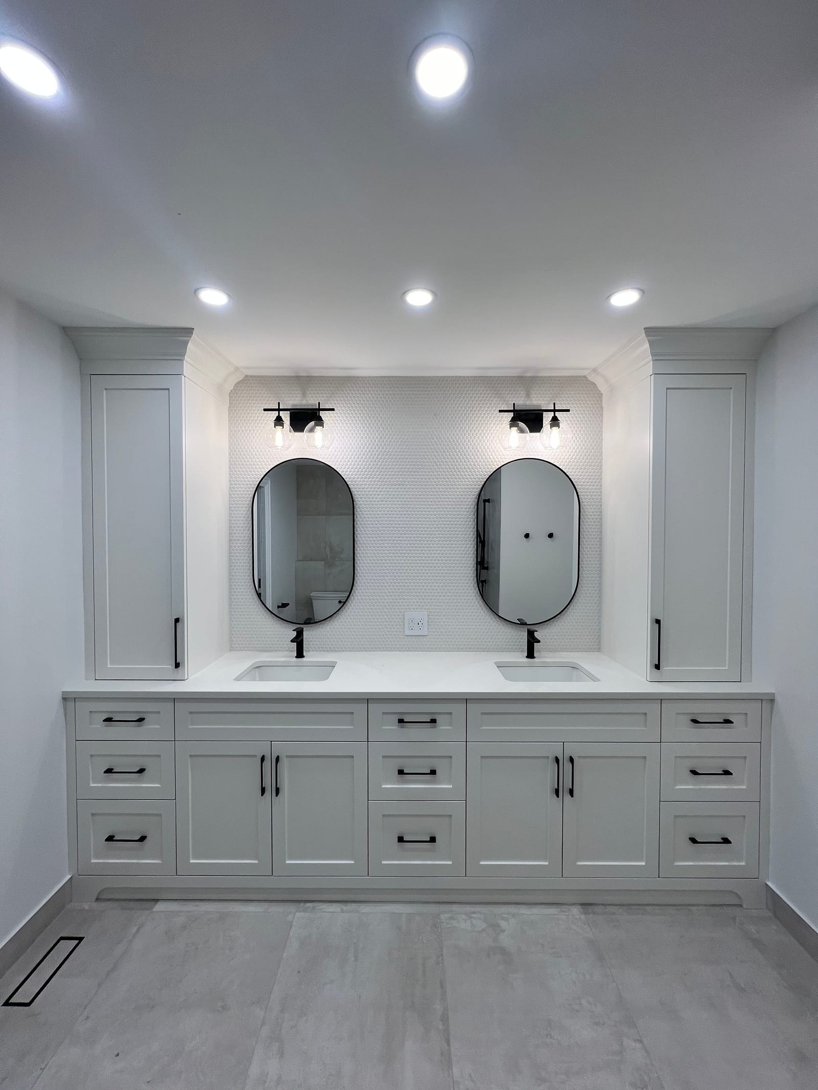 A white double vanity with two pill-shaped black mirrors, linen towers and a large-format grey floor
