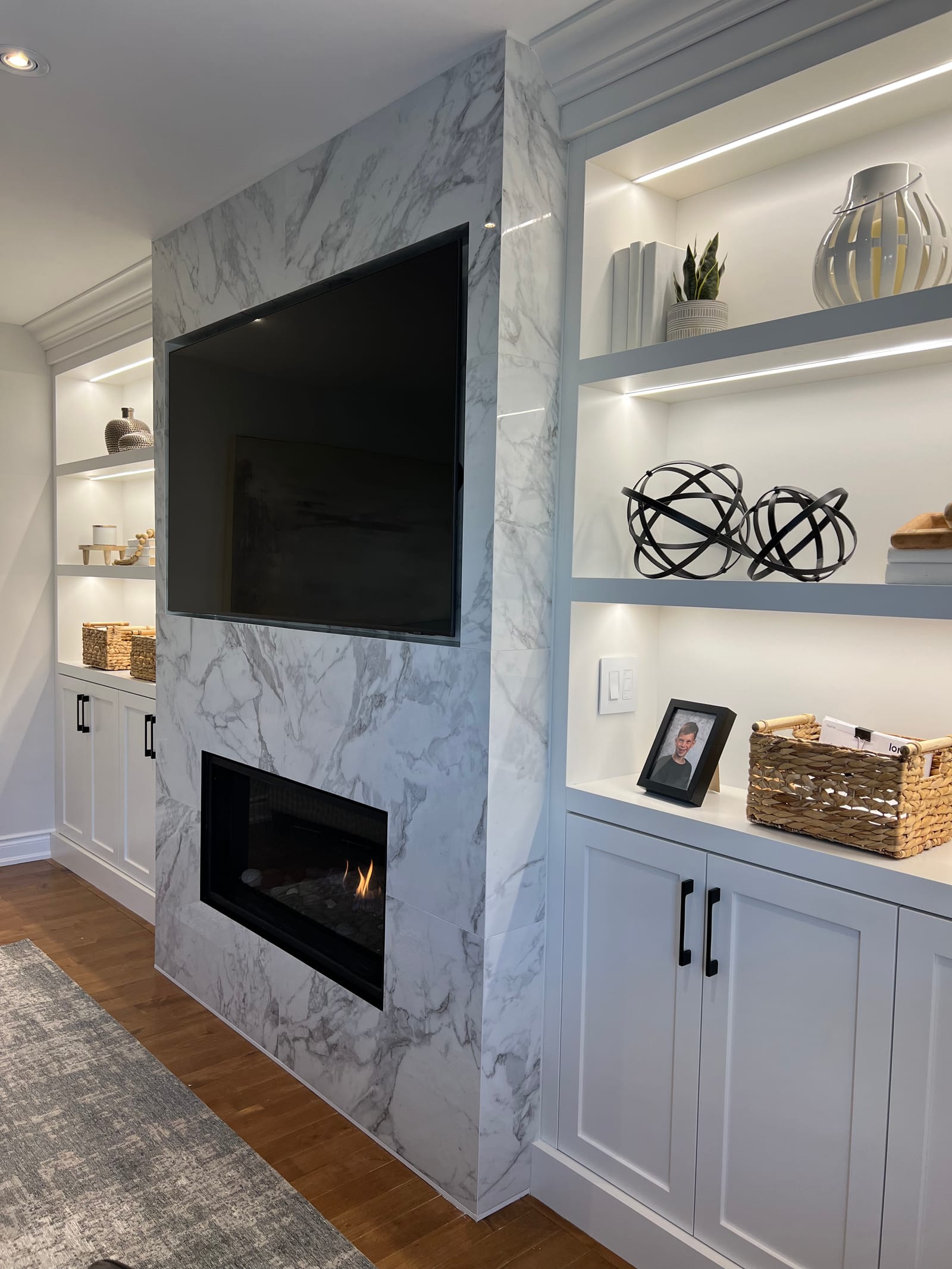 A marble-look fireplace column with a linear fireplace and TV, beside lit built-in shelves
