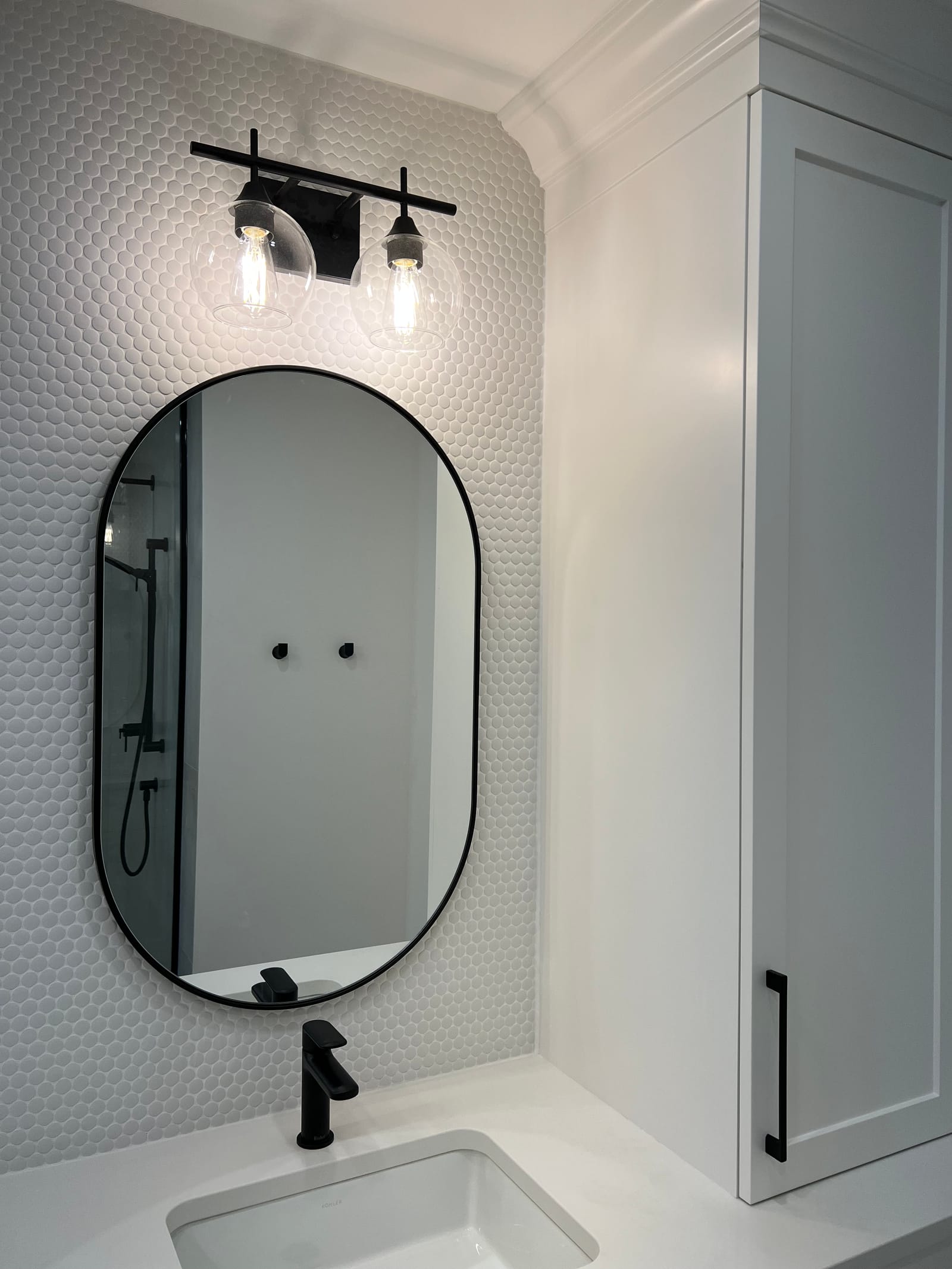 A pill-shaped black mirror over a white sink against white penny tile, with a two-globe light