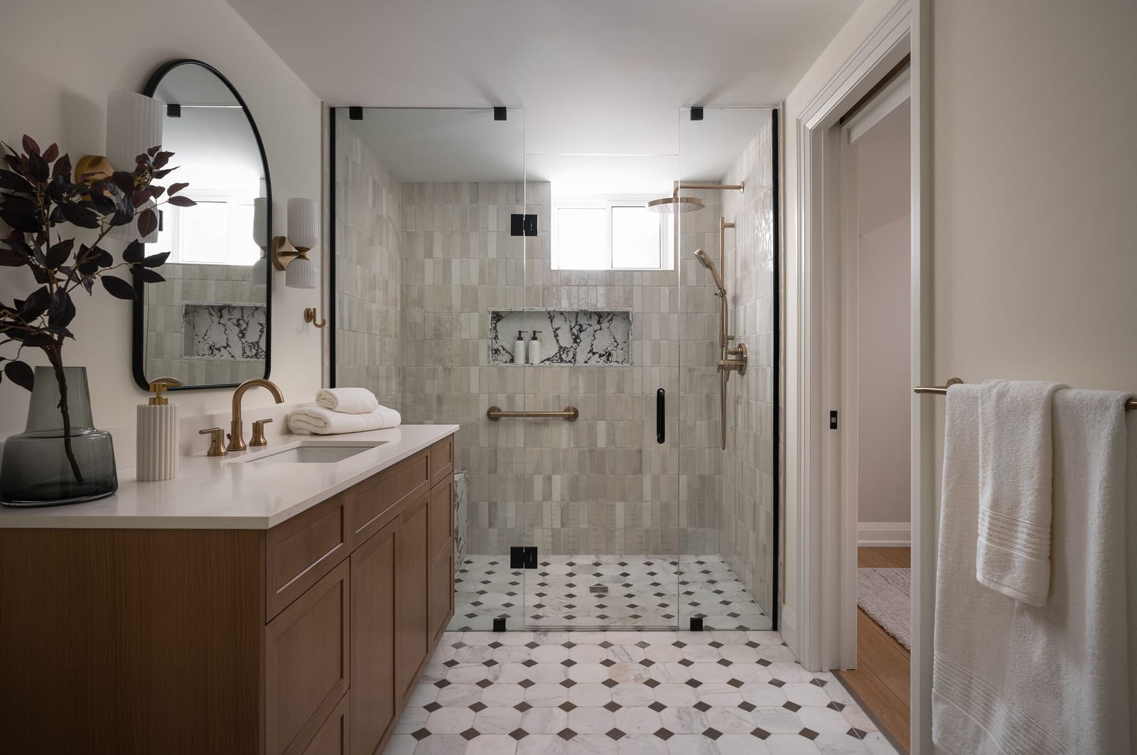 A basement bathroom with an oak vanity, an arched black mirror, a frameless glass shower with handmade-look tile and a black and white octagon floor