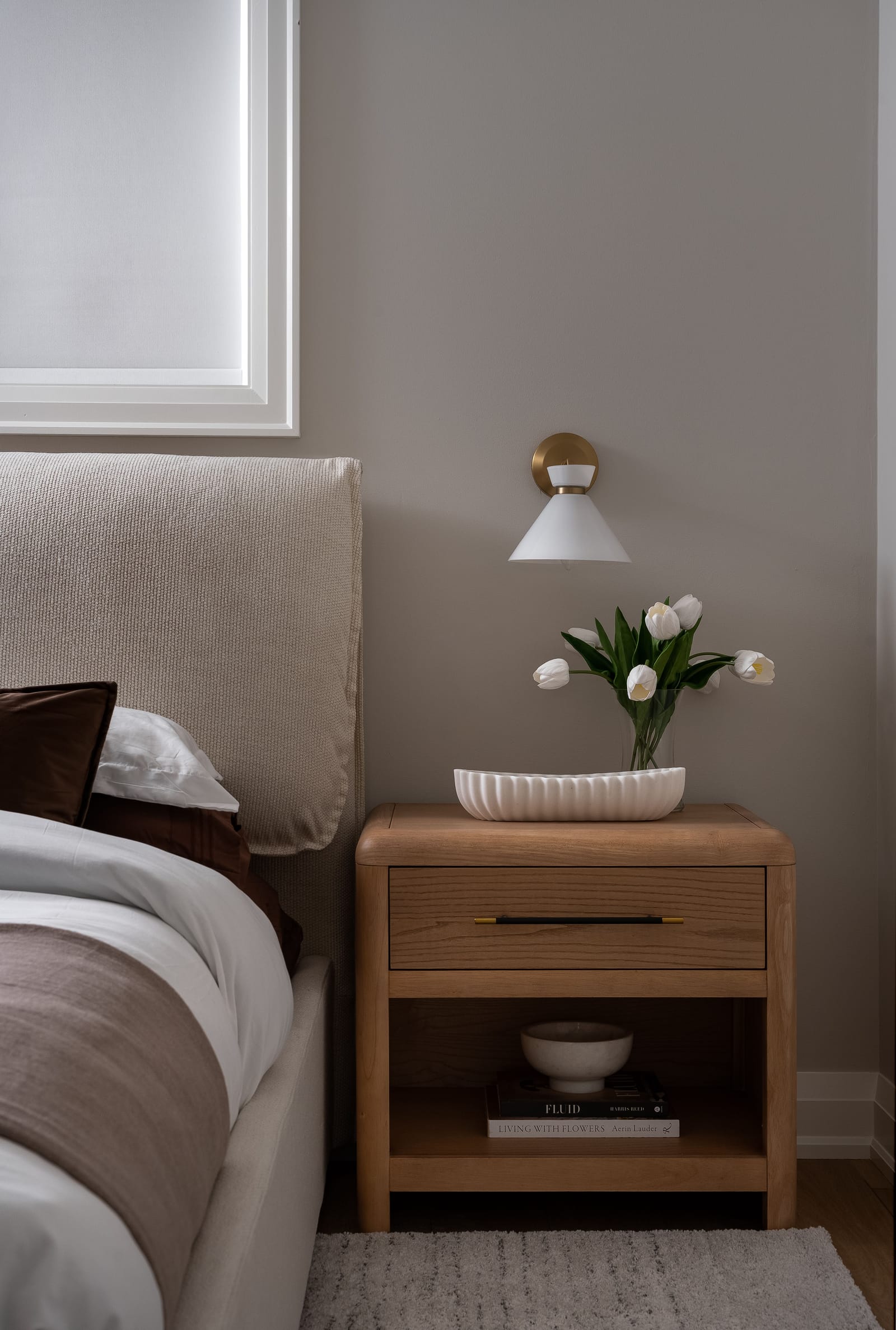 A basement bedroom corner with an upholstered headboard, an oak nightstand and a white cone wall light below a frosted window