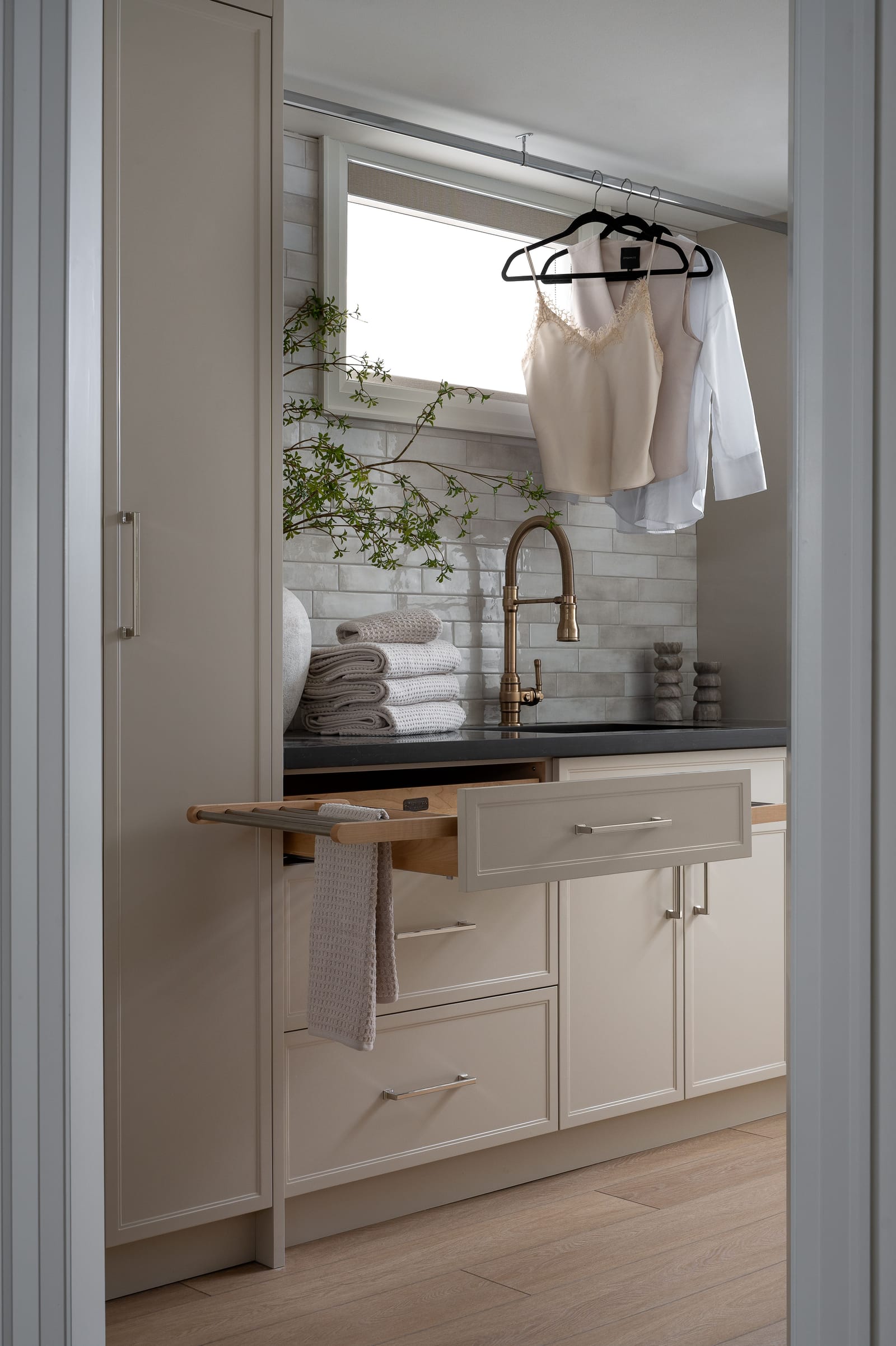 A laundry room seen through a doorway: a pull-out folding shelf, a deep sink with a brass faucet, a hanging rail and a stacked tile backsplash