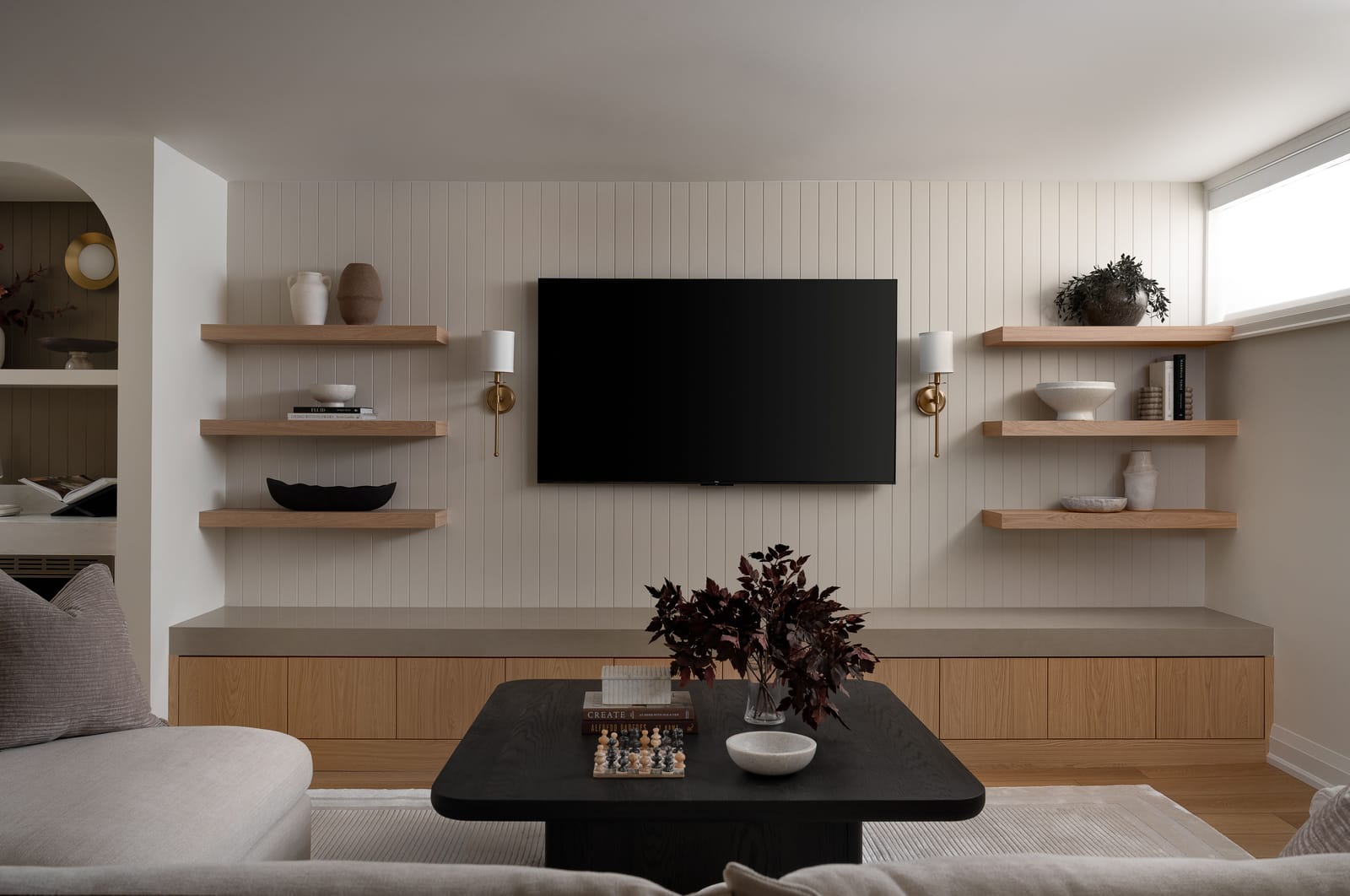 A basement rec room with a vertical-panelled media wall, oak floating shelves with brass sconces, a long oak and stone ledge and a black coffee table