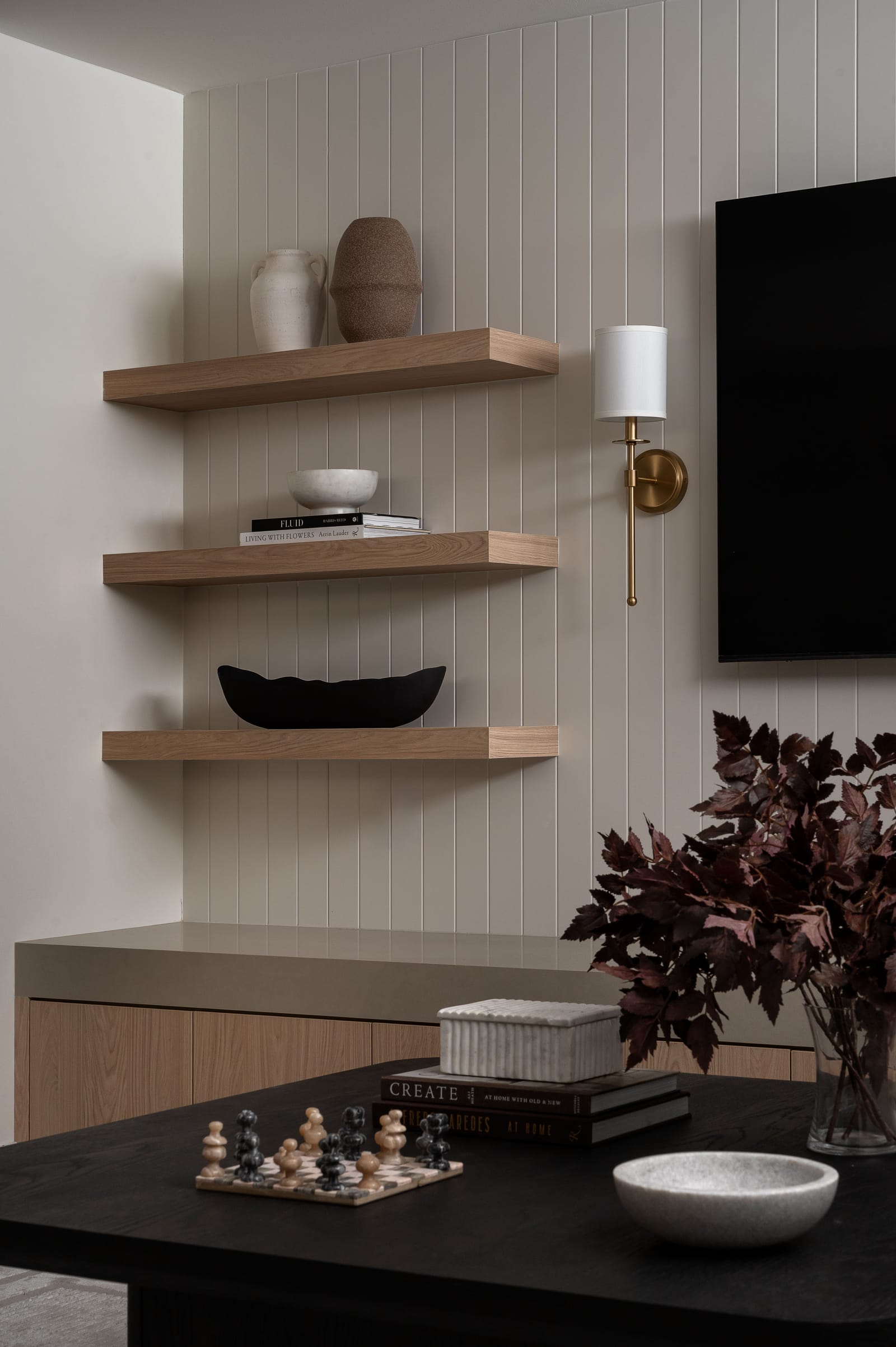 Oak floating shelves with pottery and books against white vertical panelling, lit by a brass and linen wall sconce