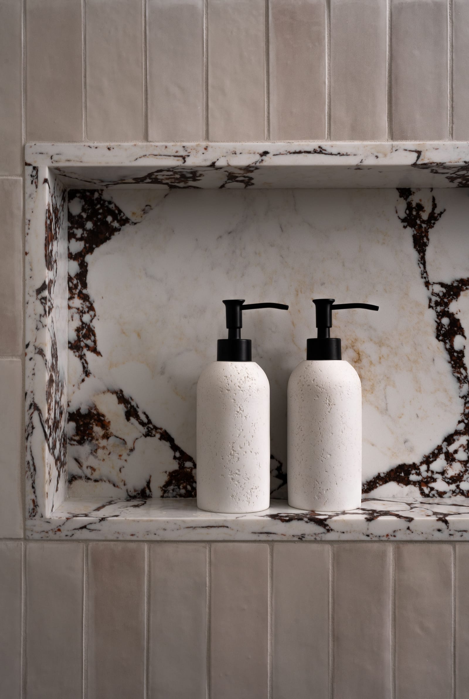 Close view of a shower niche lined with white stone with dark red and black veining, set in glossy taupe vertical tile