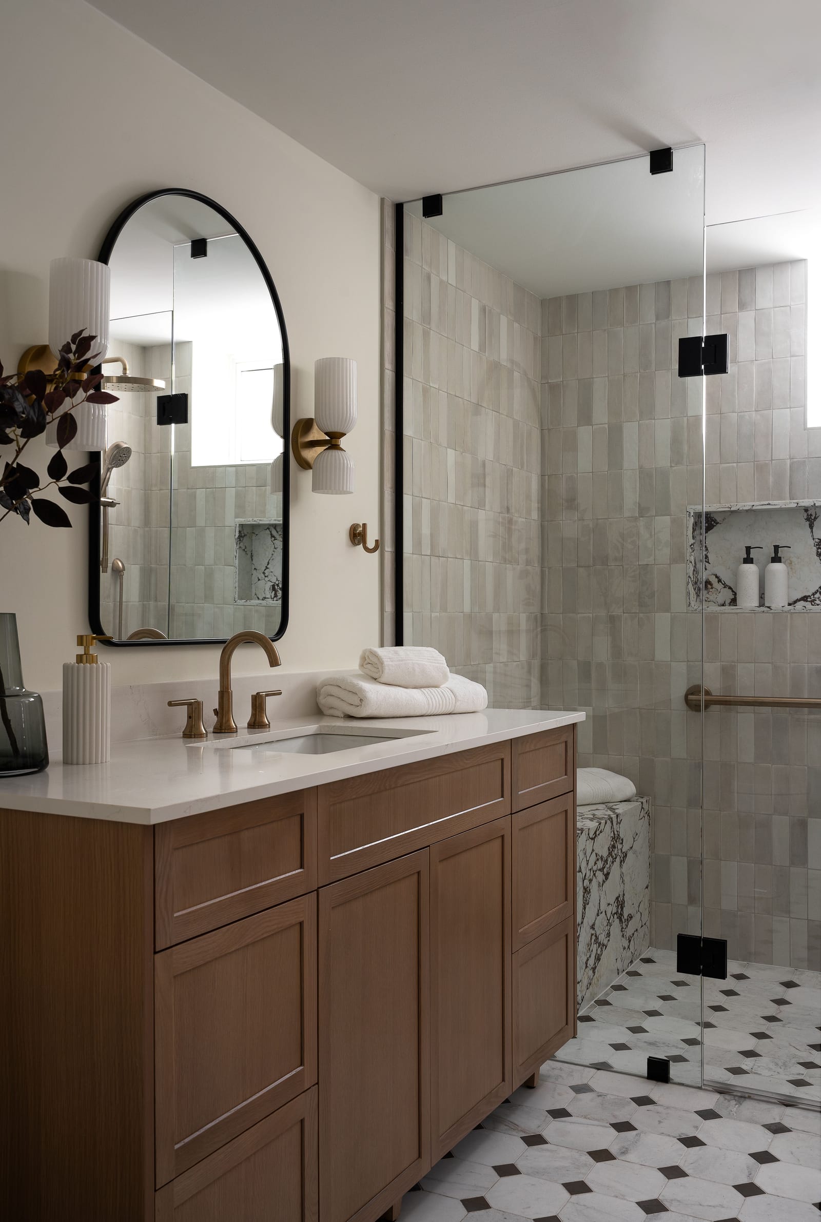 An oak shaker vanity with brass taps beside a glass shower whose niche and bench are faced in dark-veined stone