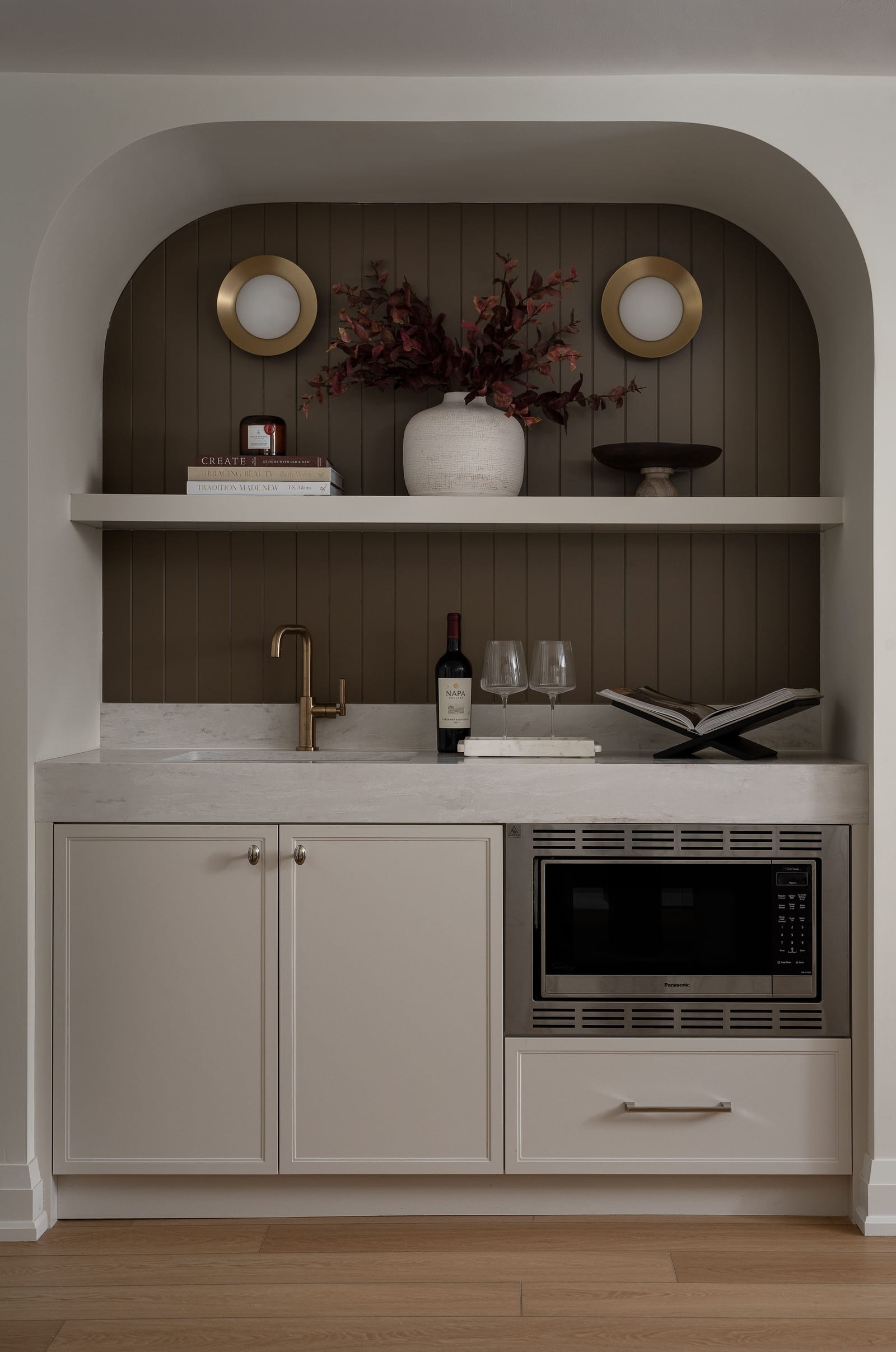 Straight-on view of the arched wet bar: dark panelled back, one shelf, two brass-rimmed round sconces, a brass faucet and cream lower cabinets