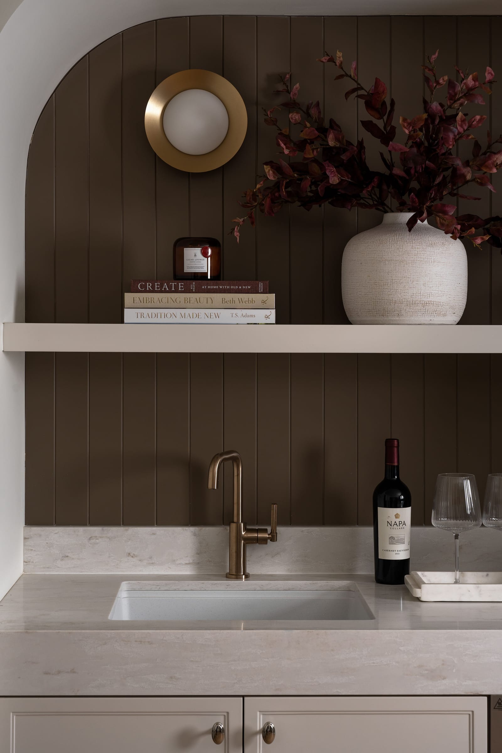 Close view of the wet bar sink with a brass faucet, a bottle of wine and glasses on a pale stone counter under the curved arch