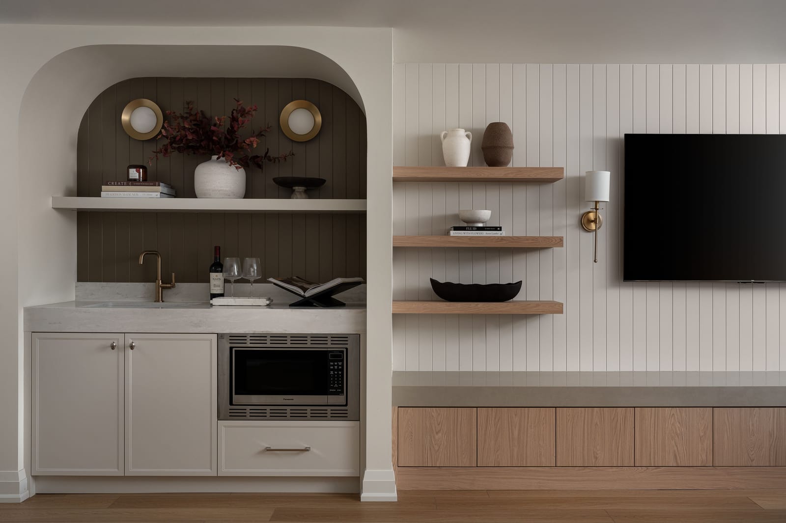 An arched alcove wet bar with dark brown panelling, a stone counter, a sink and a built-in microwave, beside the panelled TV wall
