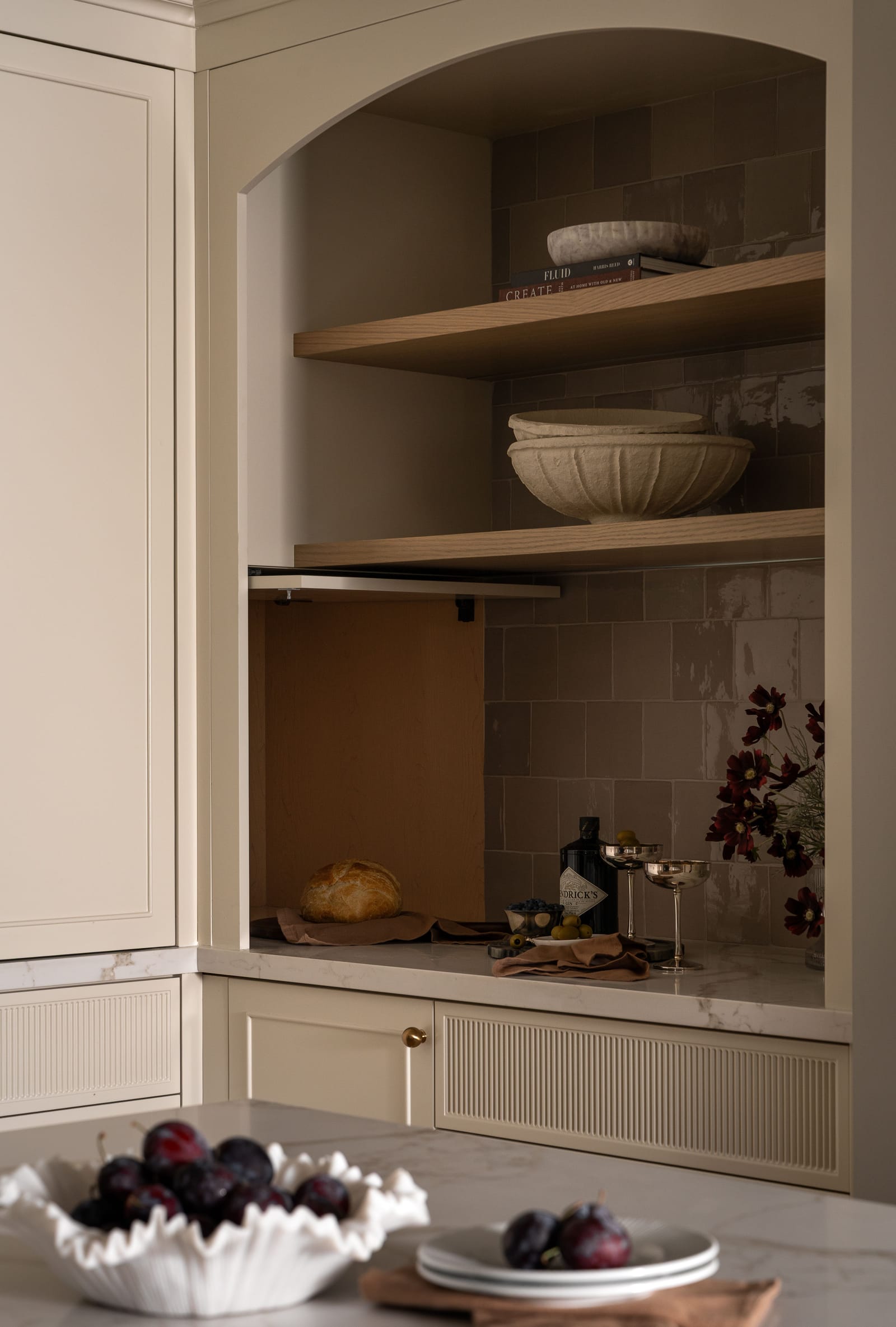 An arched niche with two oak shelves over taupe tile, and a lift-up door raised to show a wood-lined cubby with a loaf of bread inside, above cream cabinets with reeded panels