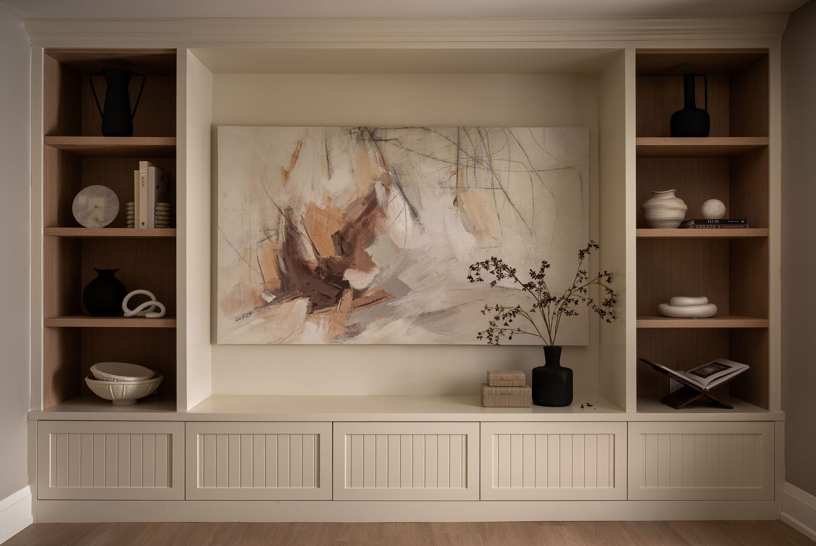 A cream built-in wall with oak-lined open shelves on each side, a long low cabinet with vertical-grooved doors and an abstract canvas in the centre recess