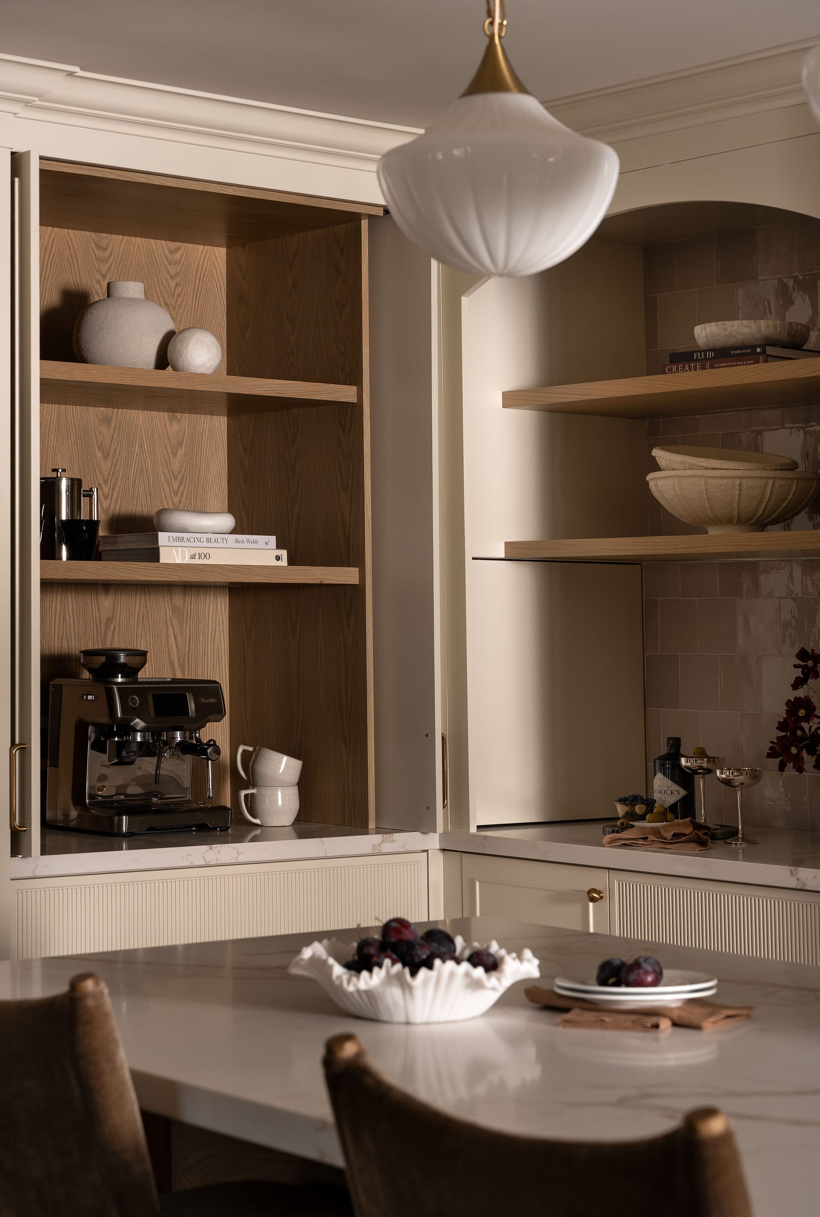 A tall cream cabinet with its doors pushed back to show oak-lined shelves, an espresso machine and mugs, beside an arched niche with oak shelves