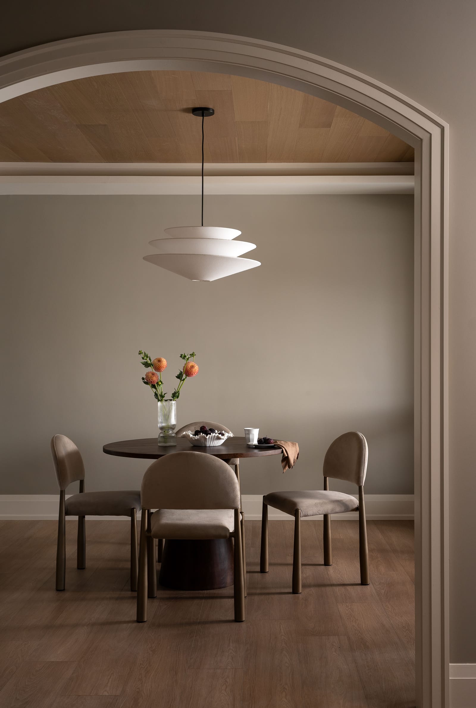 A dining room seen through a wide cased arch: an inset wood-panelled ceiling, a layered white pendant, a round dark wood table and four taupe upholstered chairs