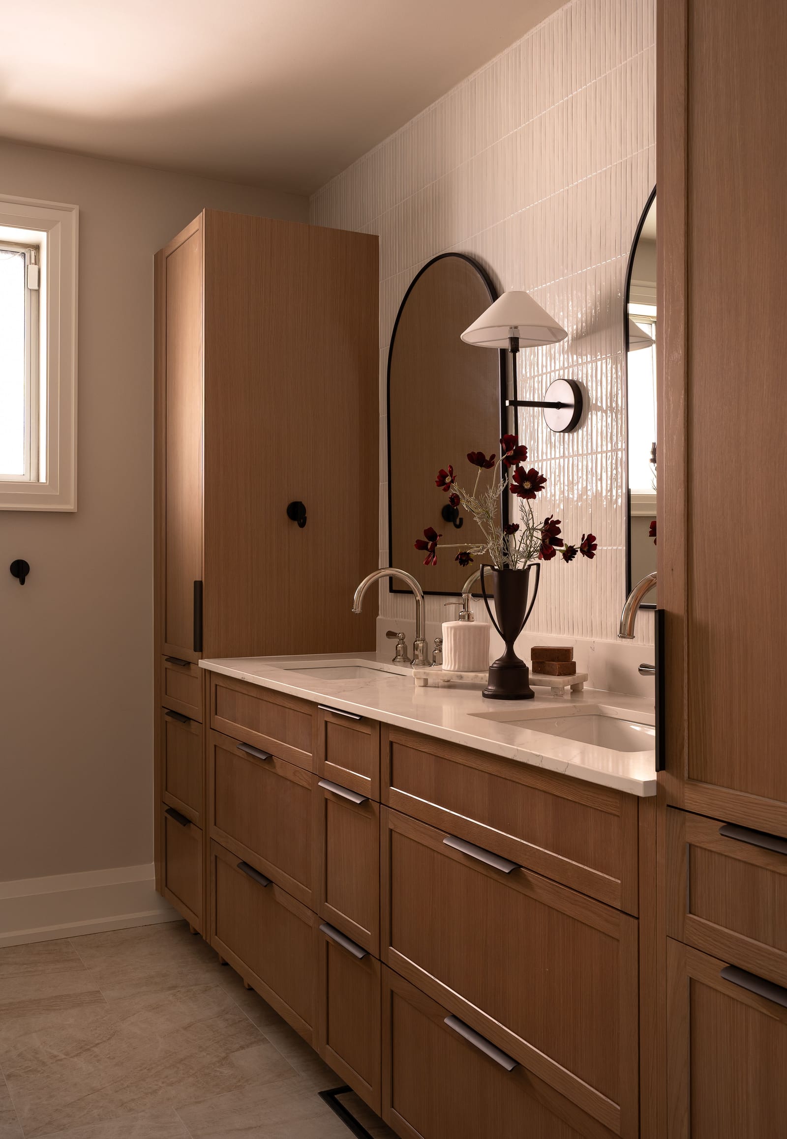 A bathroom with a long oak double vanity, tall oak linen cabinets at each end, arched black-framed mirrors, polished nickel taps and glossy white vertical tile