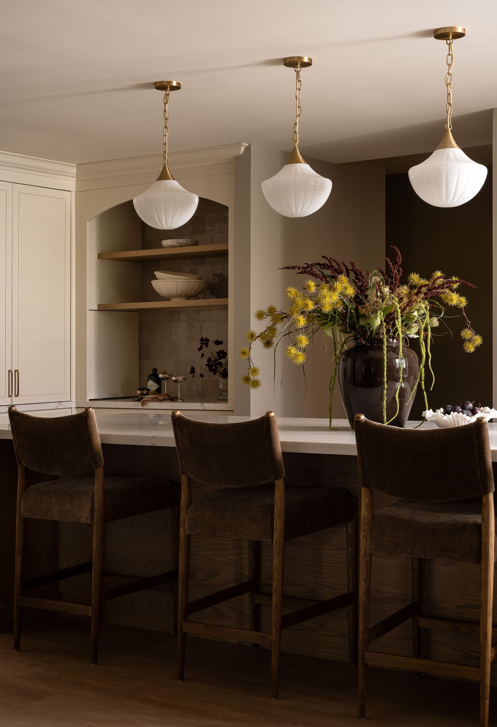 Three white fluted glass pendants on brass chains over a white veined island top, three brown velvet stools and an arched niche with oak shelves behind