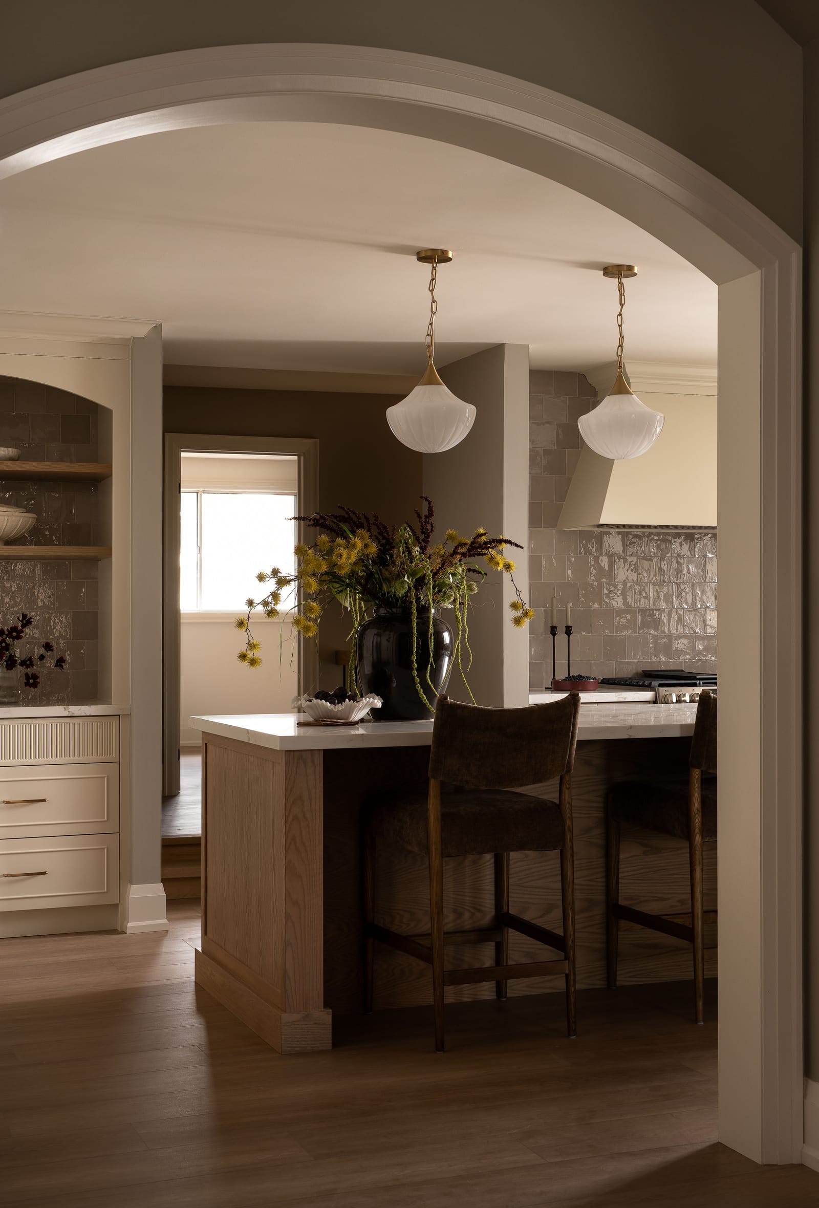 A wide cased arch framing the oak island, two velvet stools, the tiled range wall and a doorway through to a bright room beyond