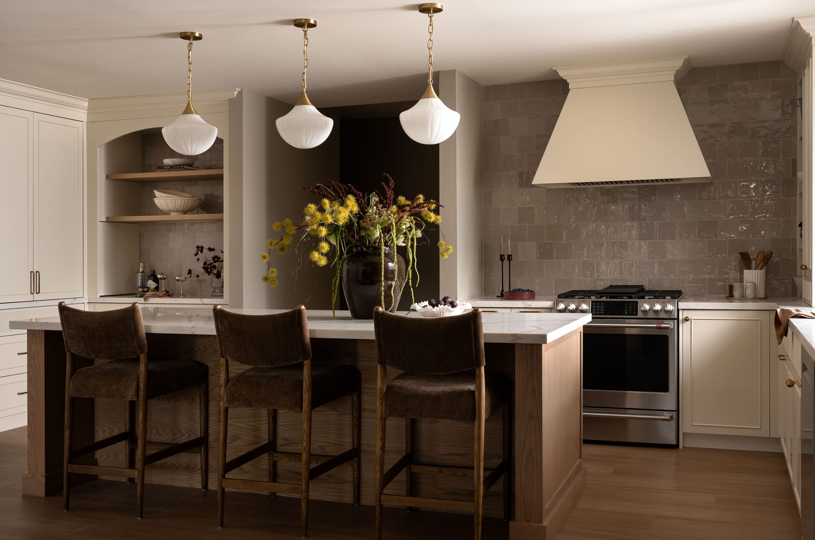 A kitchen with an oak island under three white fluted glass pendants, brown velvet stools, cream cabinets, a cream range hood and glossy taupe square tile