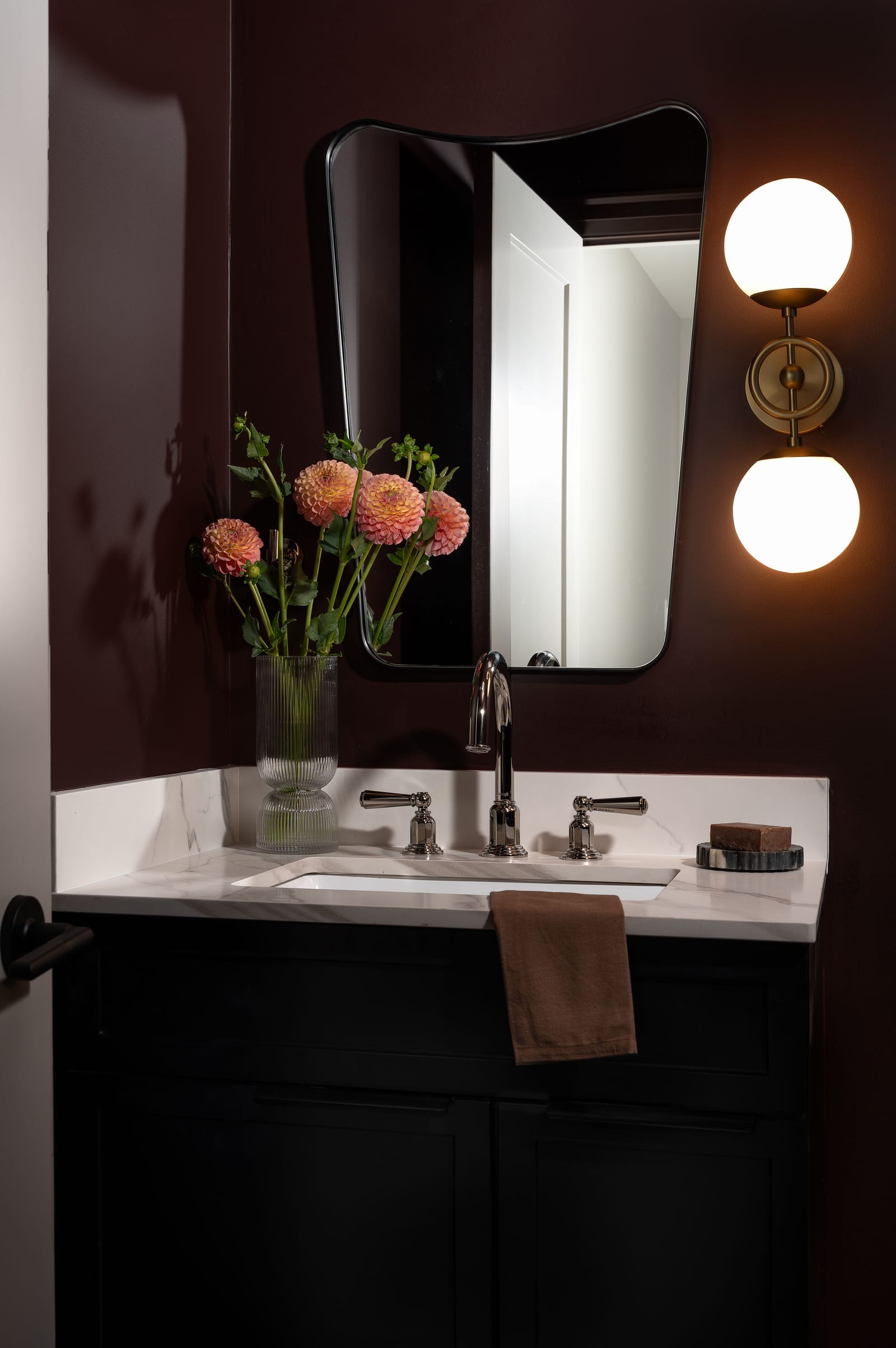 A powder room with deep plum walls, a black vanity with a white marble-look top, polished nickel lever taps, a curved black mirror and a brass two-globe sconce