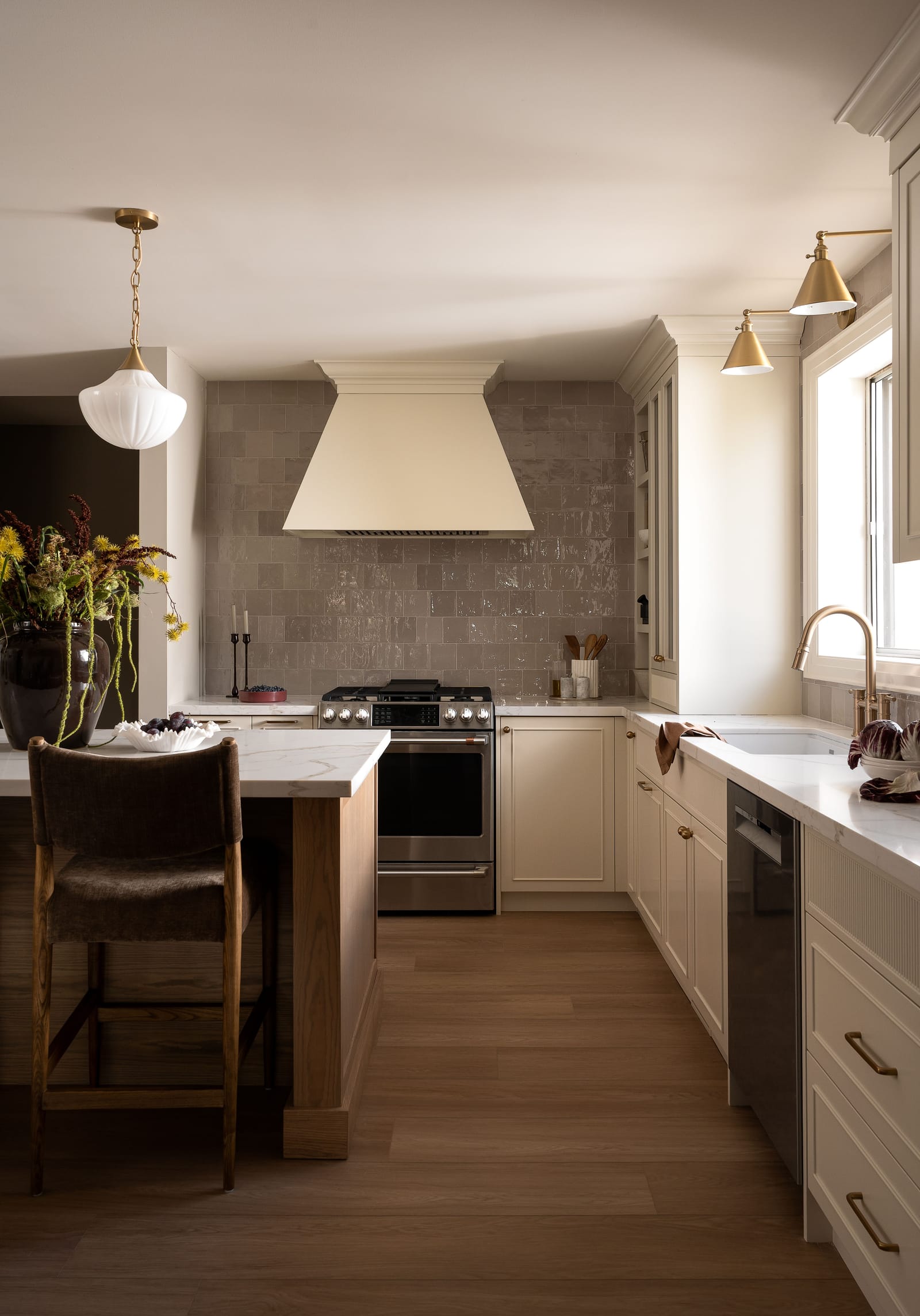 View along the kitchen to a stainless gas range under a tapered cream hood, with glossy taupe square tile to the ceiling and a wide-plank brown floor