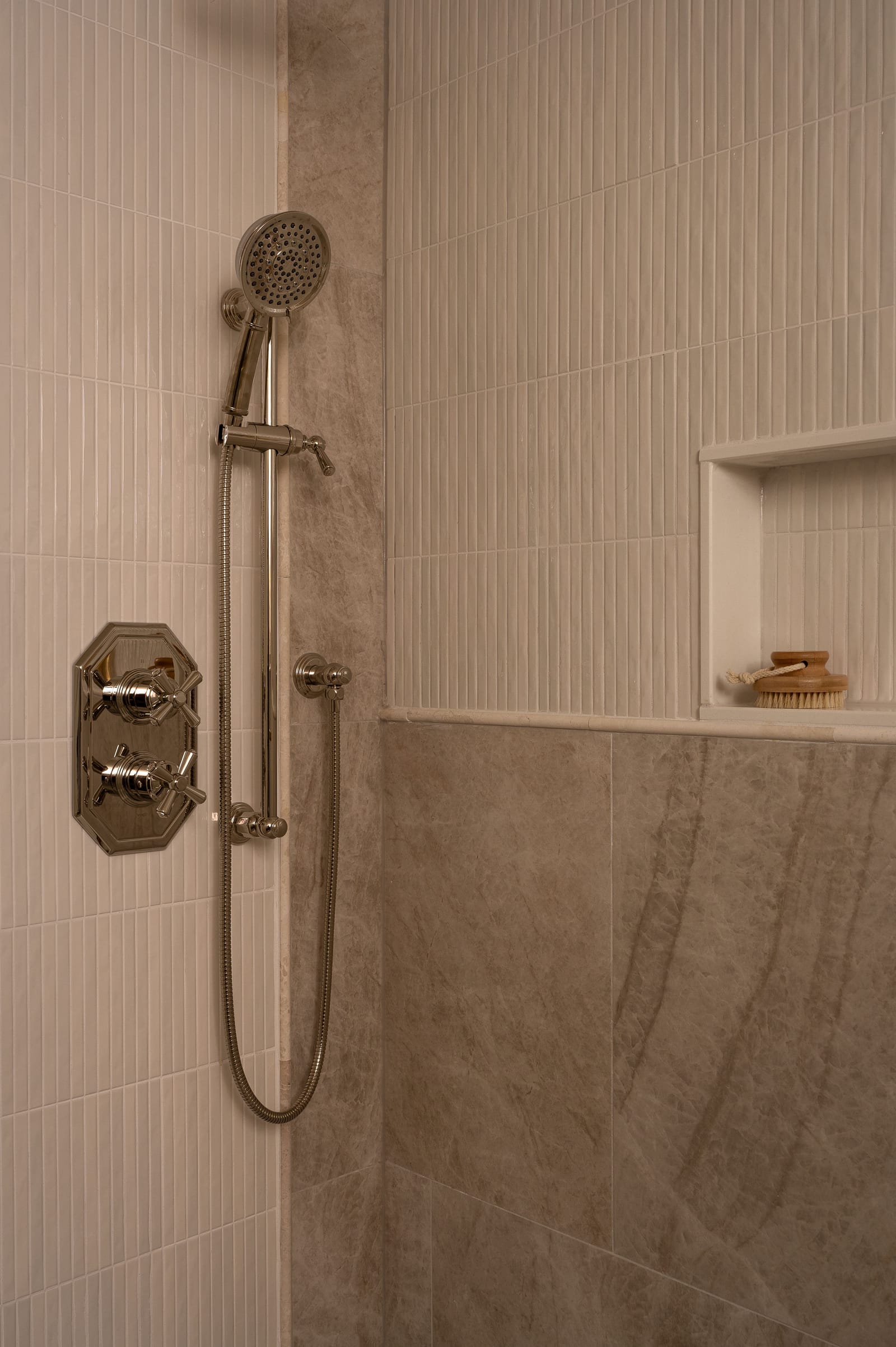A shower corner with a polished nickel valve with cross handles, a hand shower on a slide bar, white vertical stick tile, a niche and large grey veined tile below