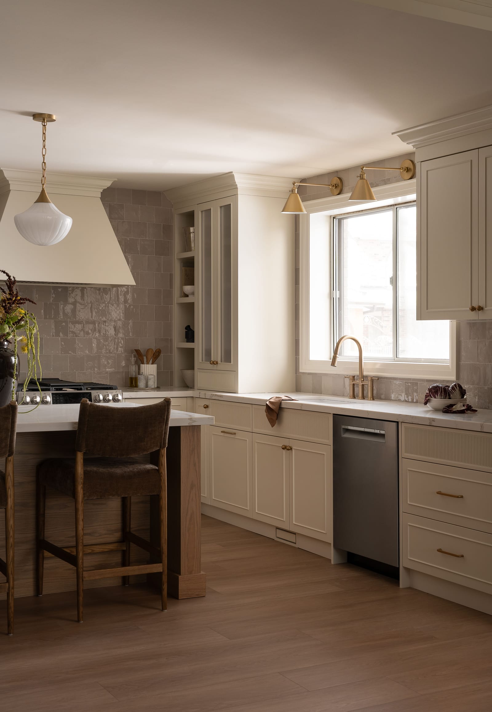 A sink under a window with a brass bridge faucet and two brass swing-arm sconces, cream cabinets with reeded drawer fronts and a glass-front upper cabinet