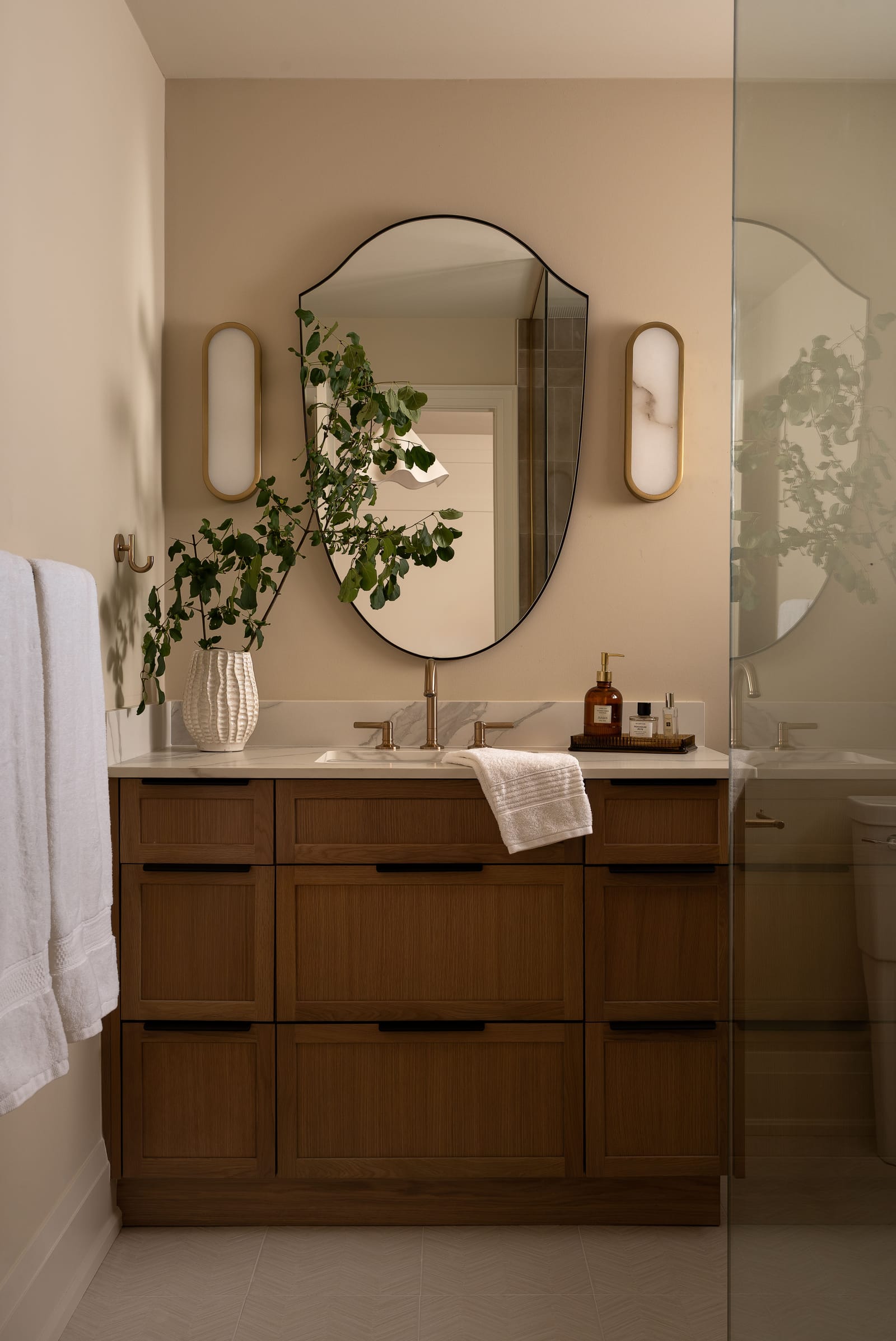 An oak shaker vanity with black pulls and a white marble-look top, a shield-shaped black mirror between two oval brass sconces, and a glass shower panel